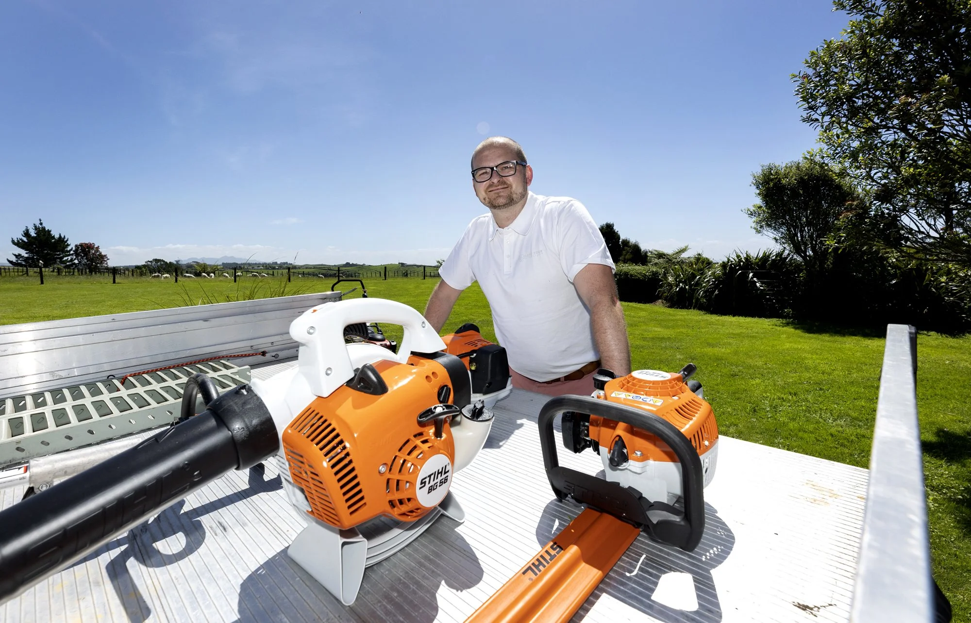 Man standing on the back of a truck with outdoor tools, including a leaf blower and chainsaw, on a bright sunny day with green grass and trees in the background.
