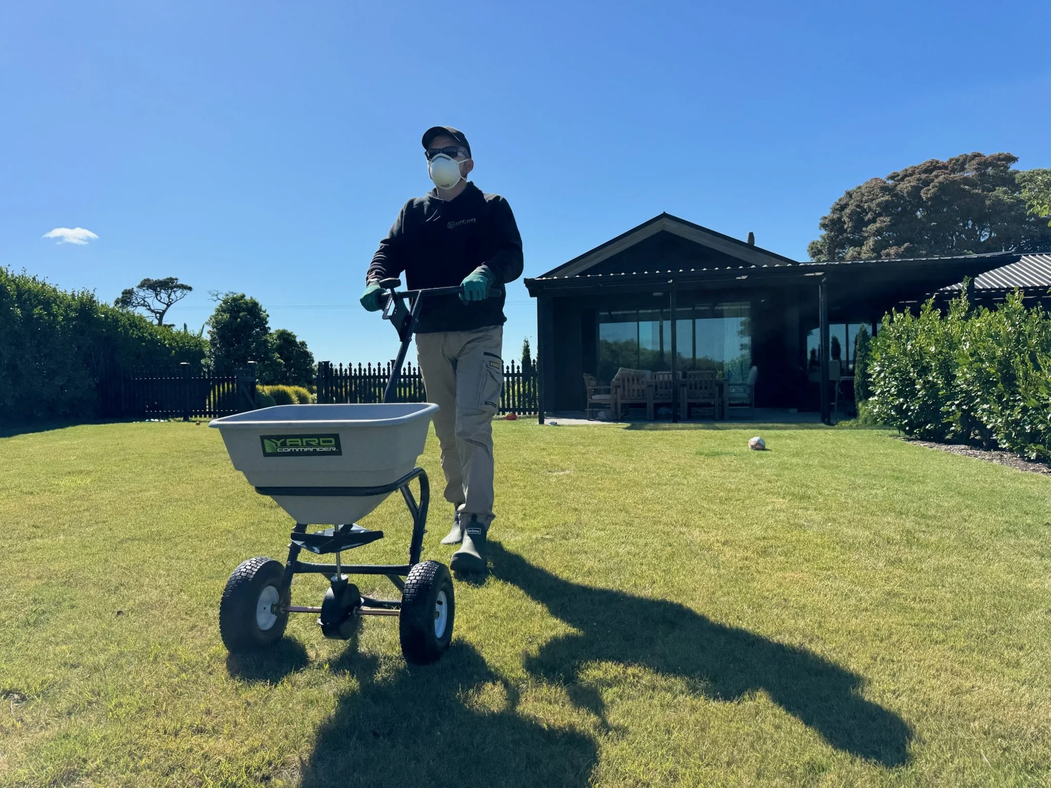 Person wearing a face mask, gloves, and sunglasses standing on a lawn, holding a garden sprayer, with a garden house and bushes in the background.