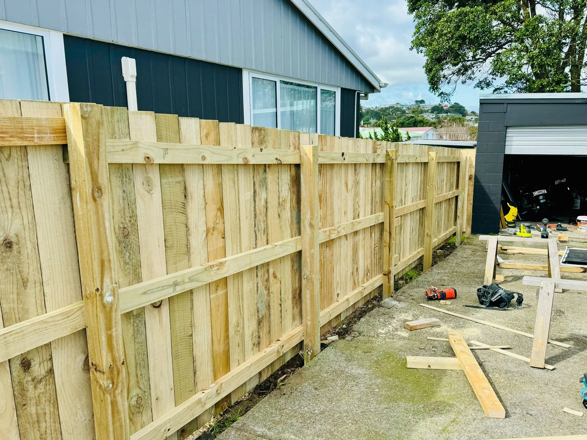 A wooden fence under construction in a backyard with tools and building materials scattered on the concrete ground.