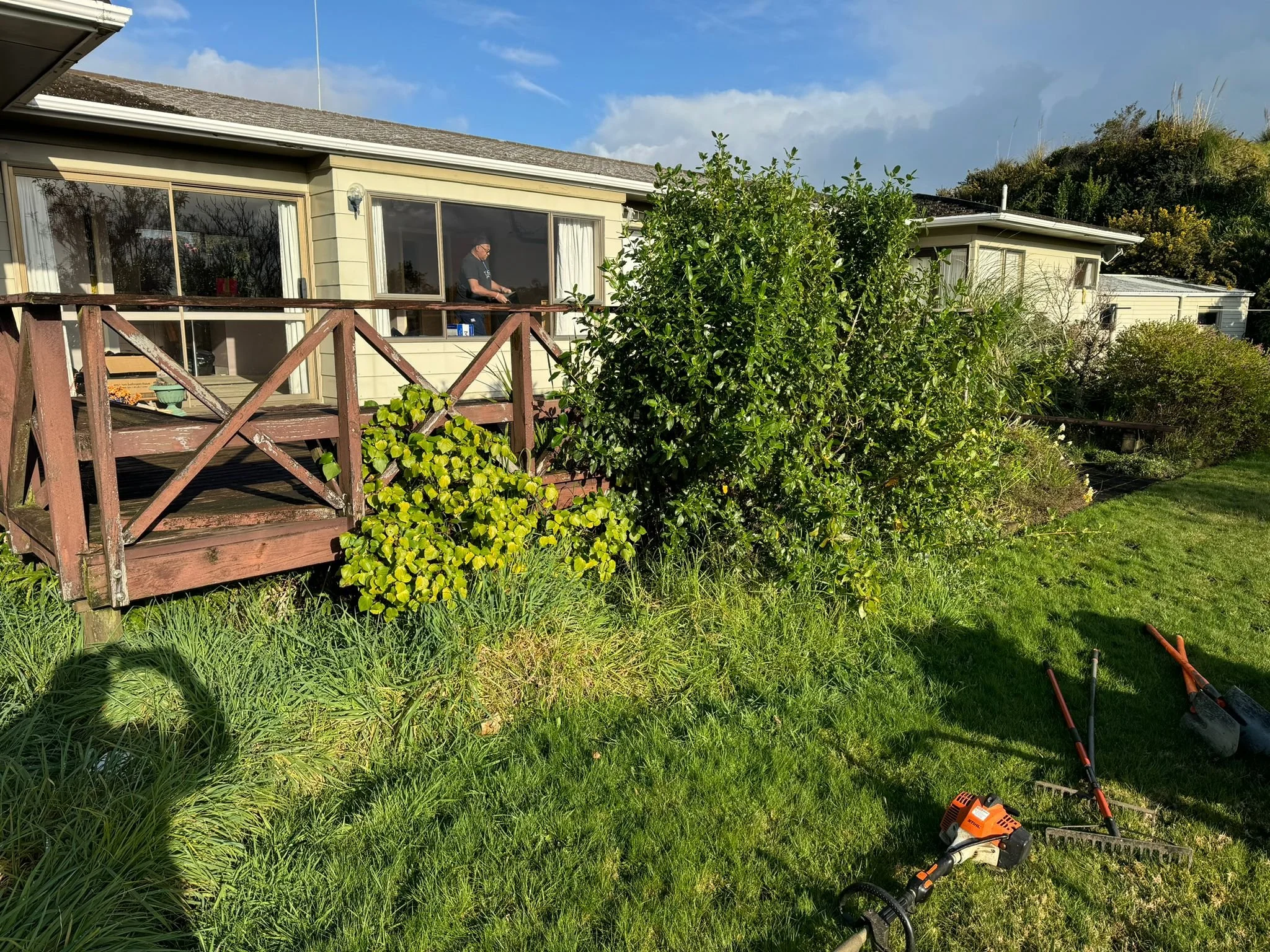 A backyard with a wooden deck attached to a house. The deck has a man working on it, and there are gardening tools, including a hedge trimmer and a rake, on the grass nearby. Bushes and grass are visible in the yard, and the house has large sliding glass doors.