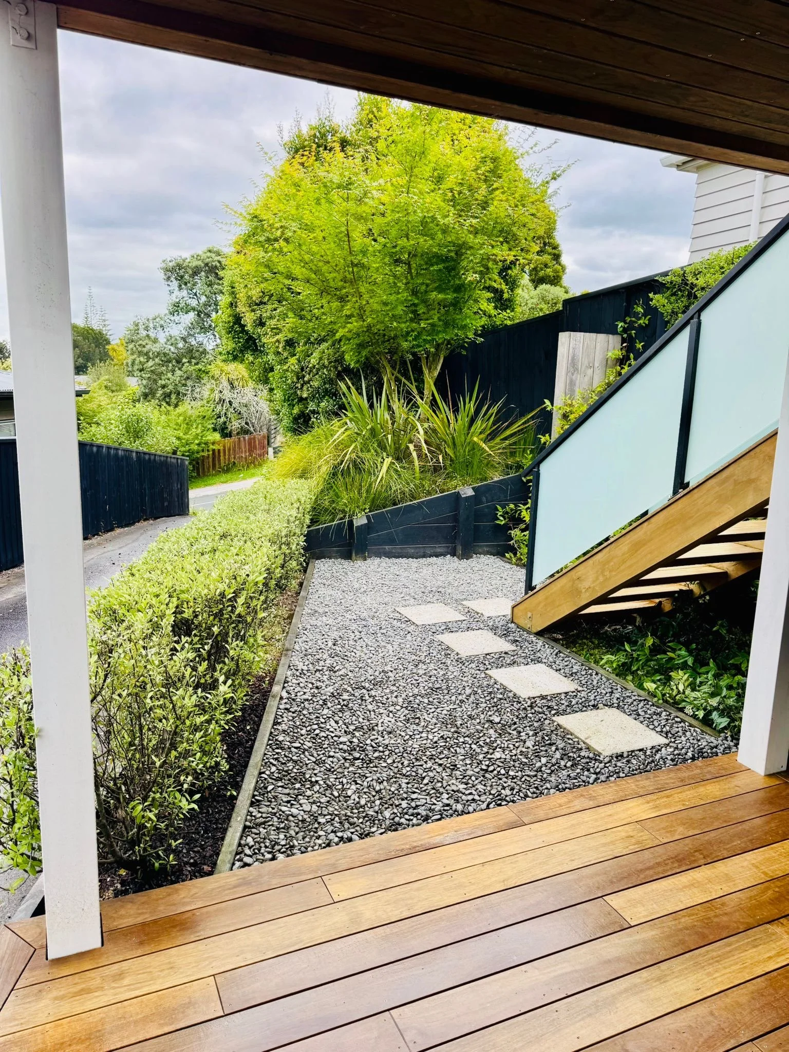 View of a backyard area with wooden deck, gravel pathway, plants, a tall tree, and a black fence.