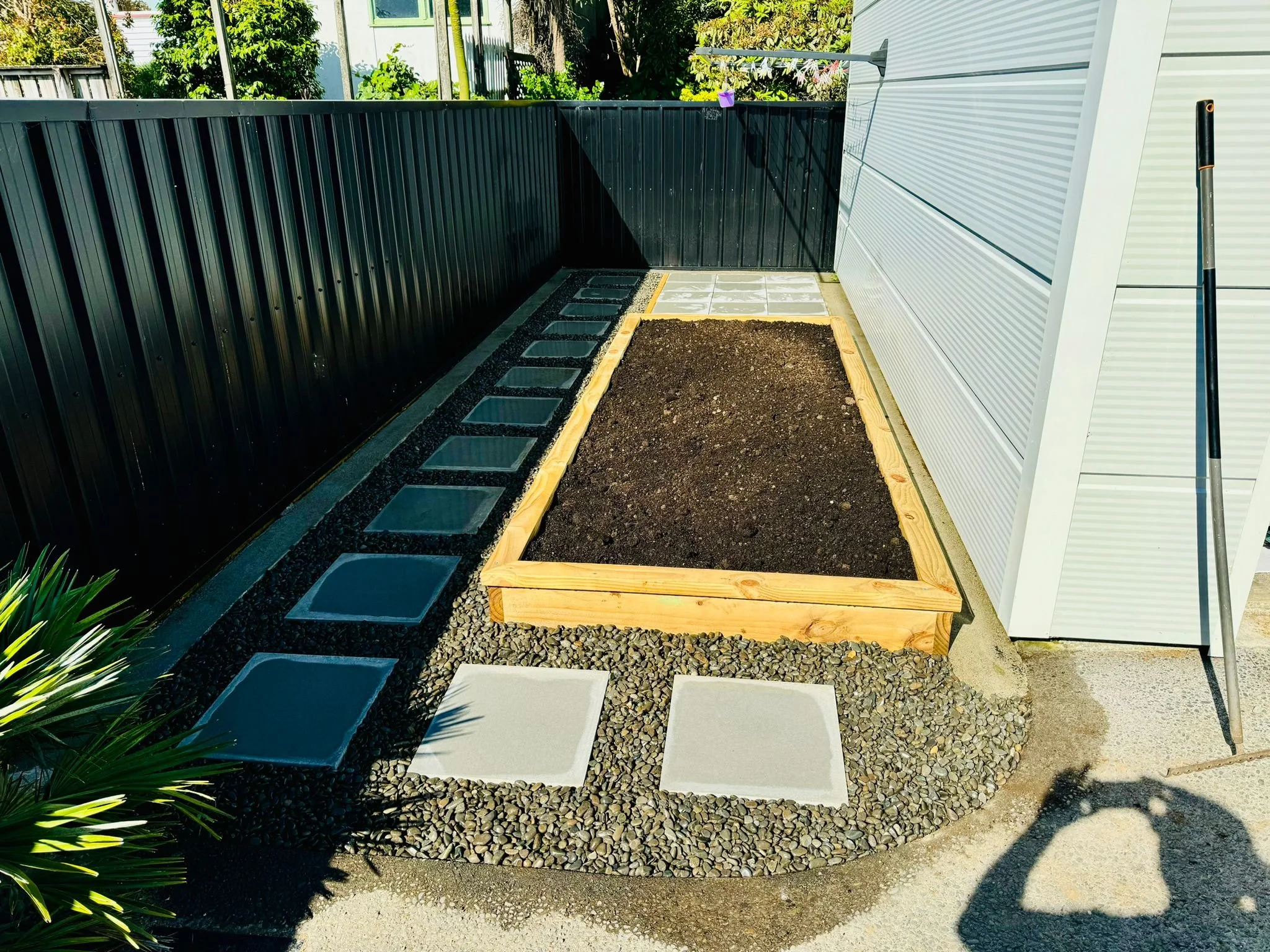 A small garden bed with dark soil bordered by wooden planks, surrounded by gravel and concrete stepping stones, with a black metal fence on one side, a white house wall on the other, and a shadow of a person taking a photo.