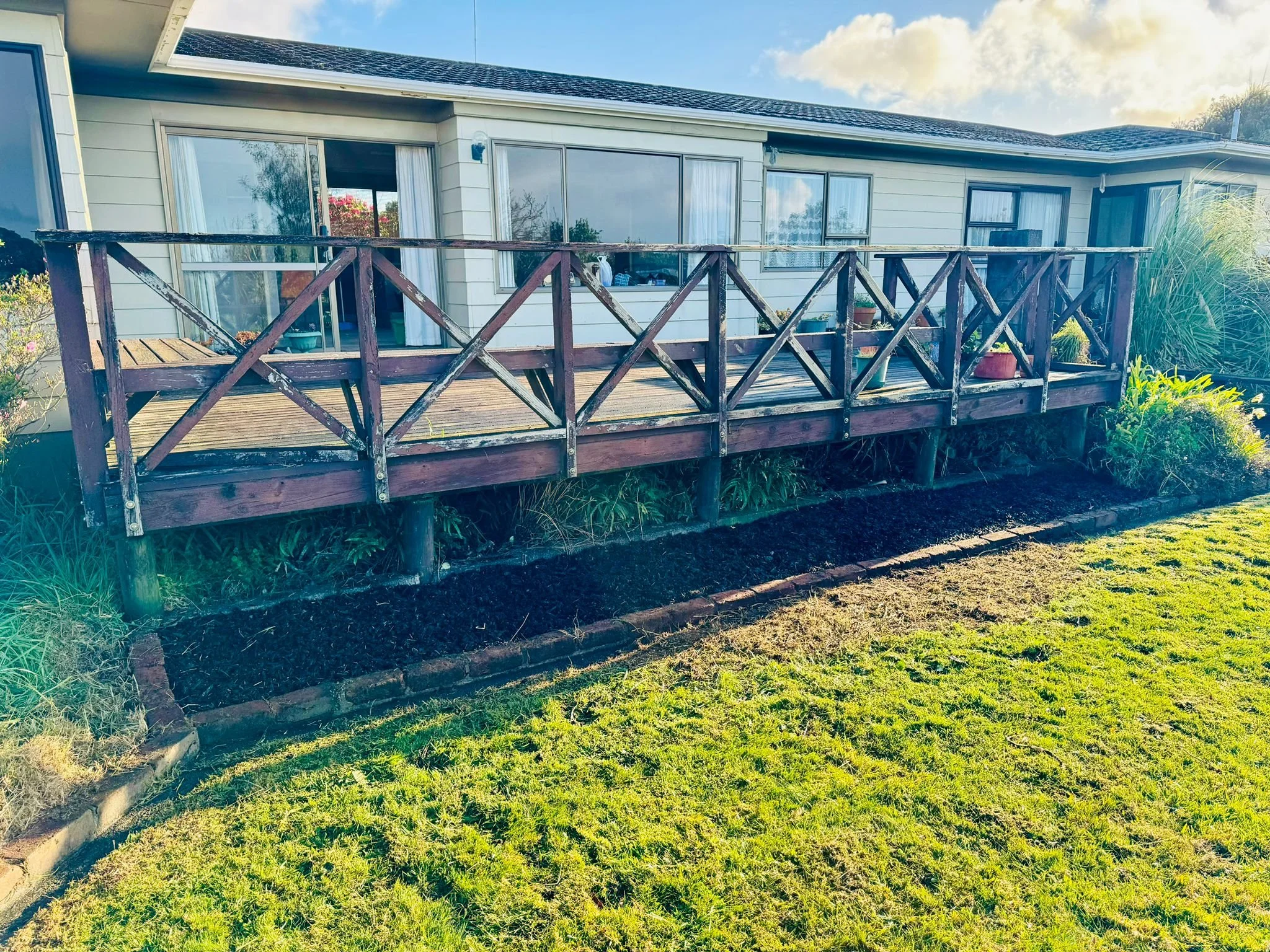 Backyard wooden deck attached to house with sliding glass doors and windows, surrounded by grass and plants.