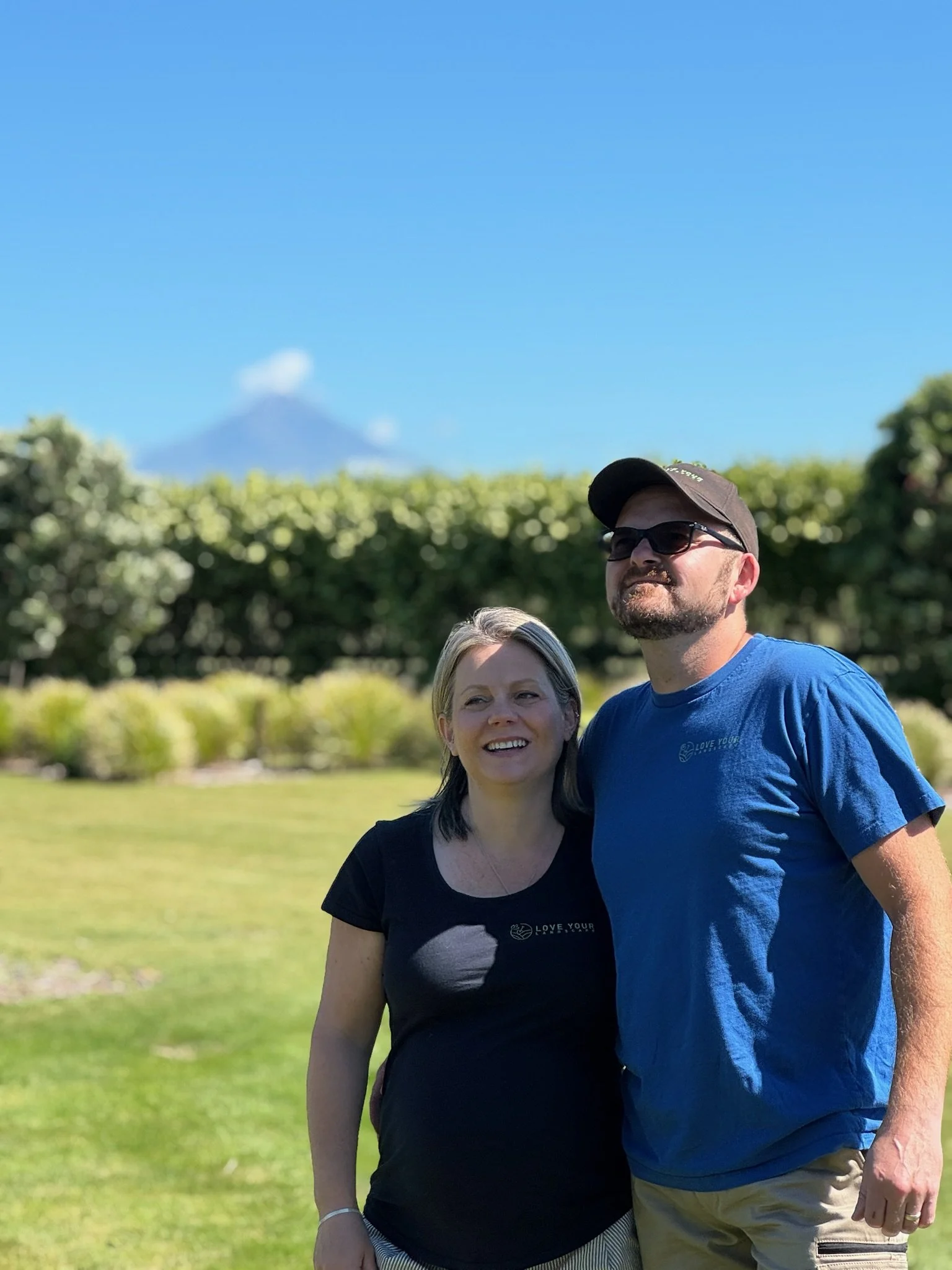 A smiling woman and a man standing outdoors on a sunny day with green grass and bushes in the background, mountain with snow on top in the distance.