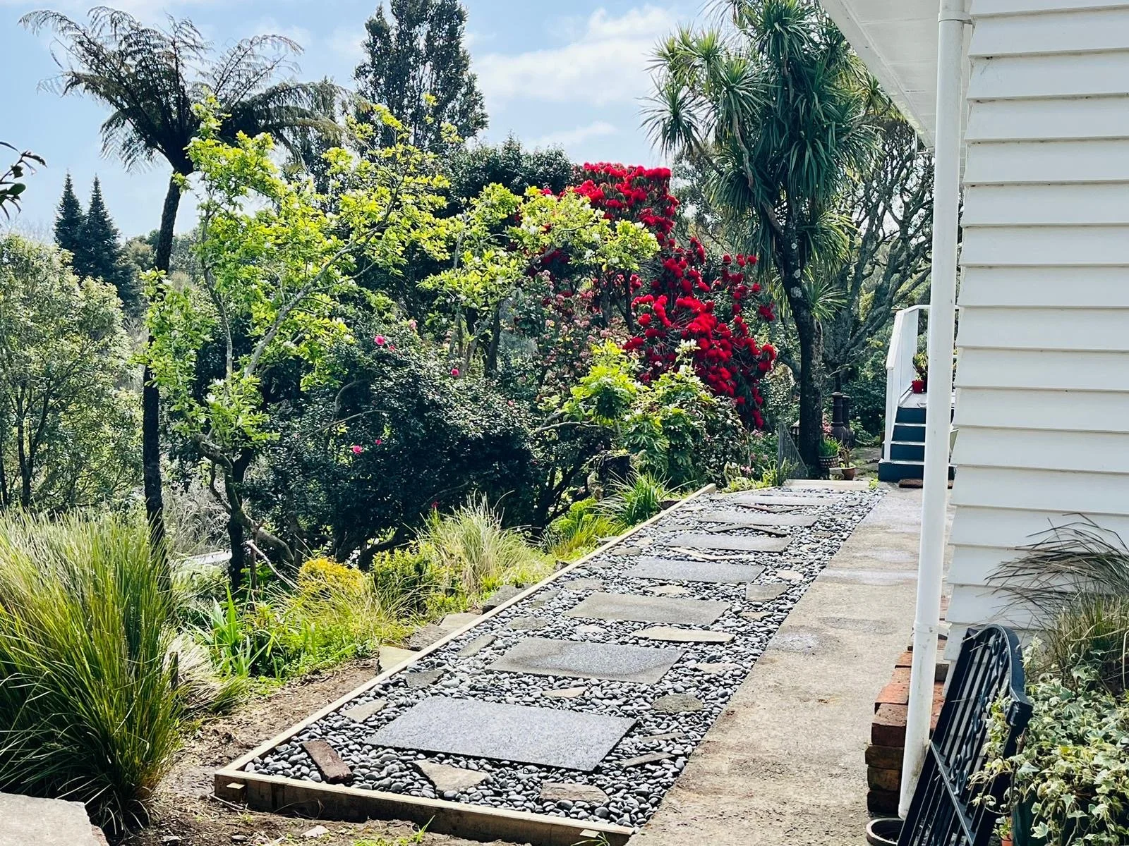 A garden pathway made of stepping stones and black gravel runs alongside a white house with a side staircase. The garden has various green plants, shrubs, and trees, including a large red-flowered shrub and a tall palm tree. The sky is partly cloudy.