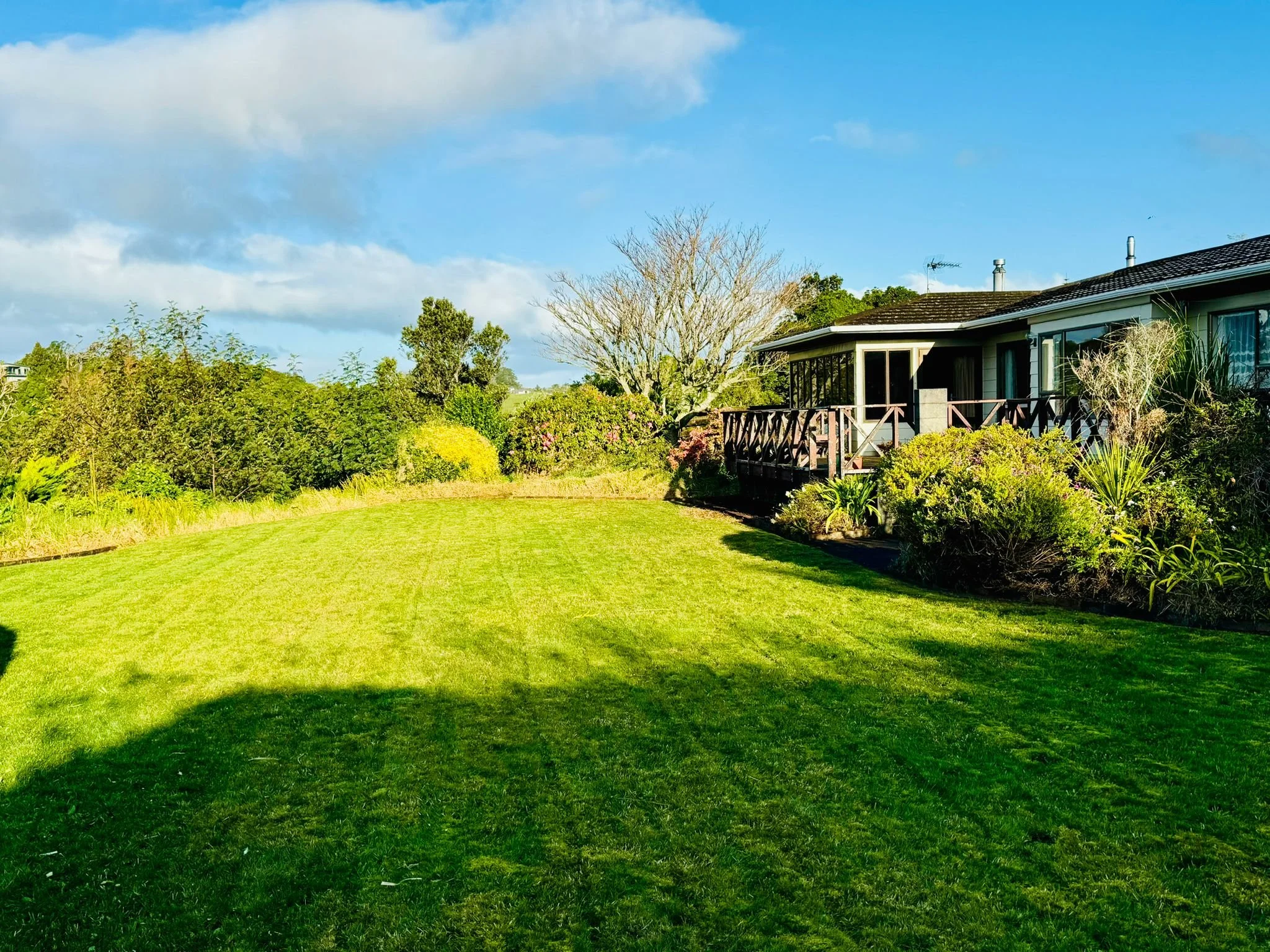 A spacious backyard with a well-maintained green lawn, a house with a wooden deck, and various trees and shrubs under a partly cloudy sky.