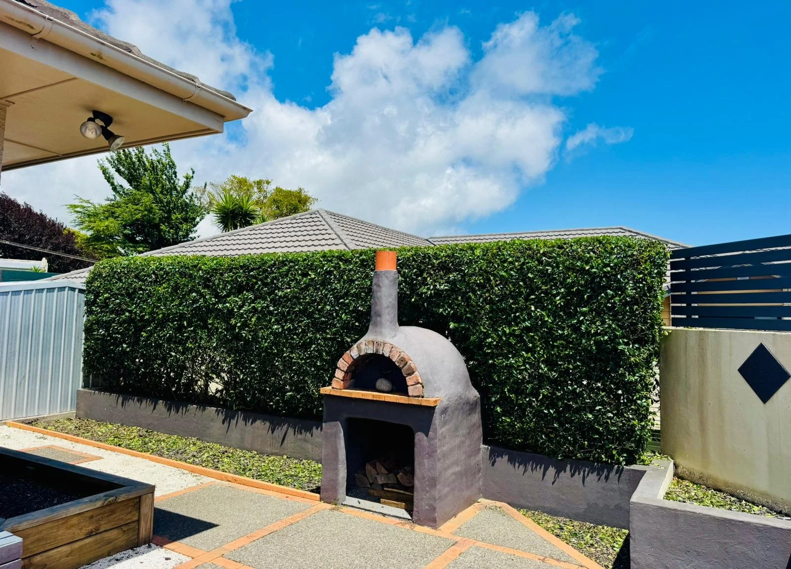 Outdoor backyard with a small brick oven, a green hedge, a concrete fence with a diamond shape, and a blue sky with clouds.