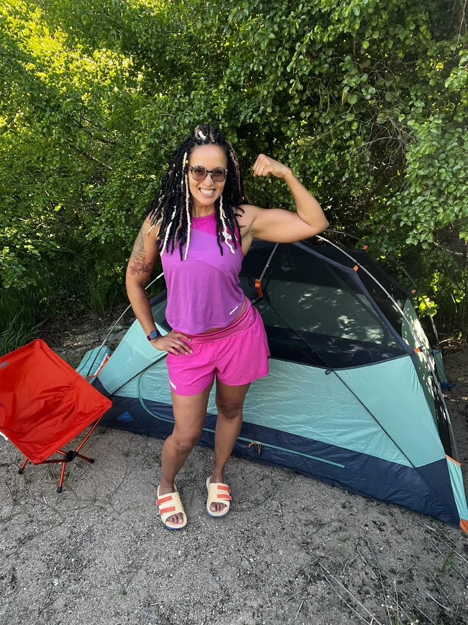 Women of color smiling and flexing in front of a tent and a camping chair.