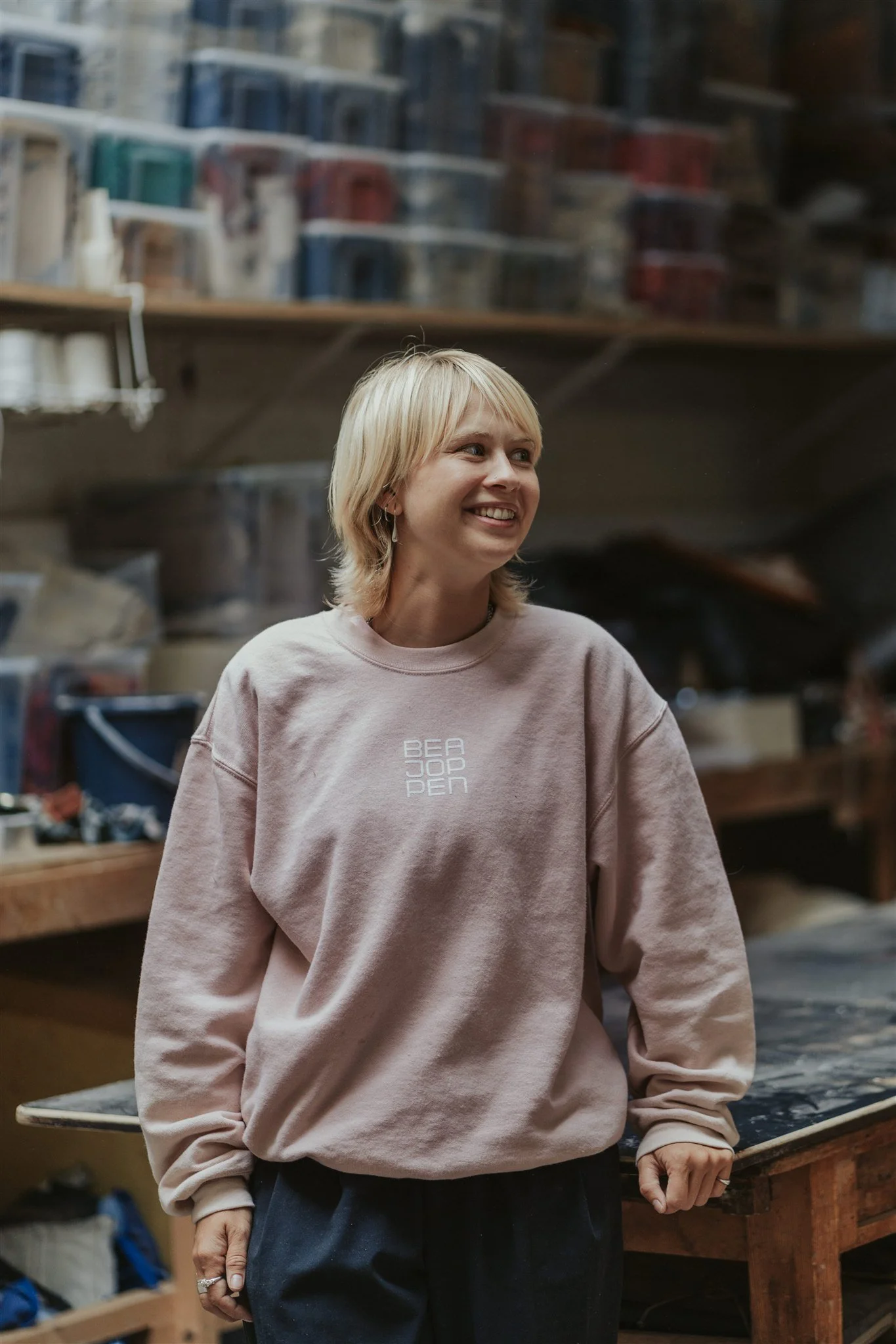 A woman with blonde hair wearing a light pink sweatshirt and black pants standing in a workshop with shelves of fabric or supplies in the background.
