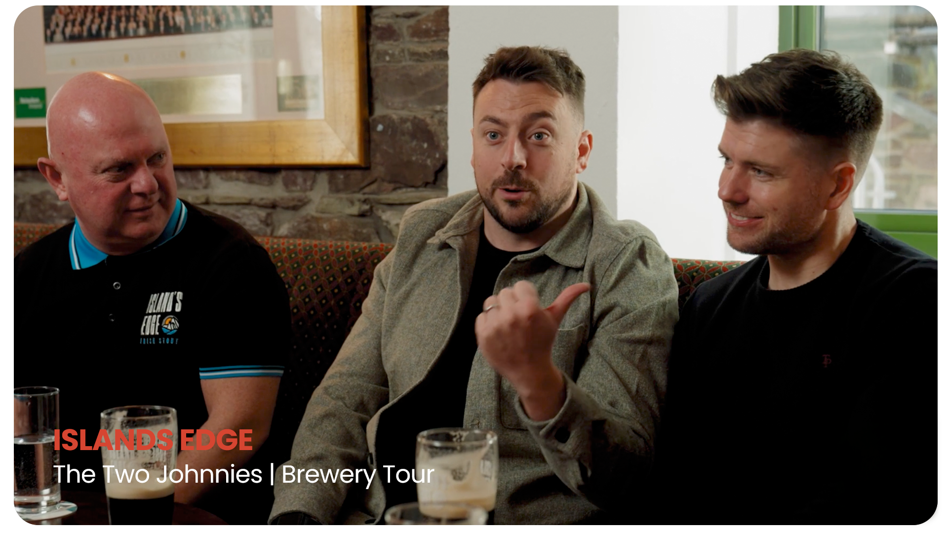 Three men sitting at a table in a brewery or pub, with drinks in front of them, engaged in conversation. The man in the middle is speaking, with a hand gesture, while the other two listen. The background shows a brick wall with a window.