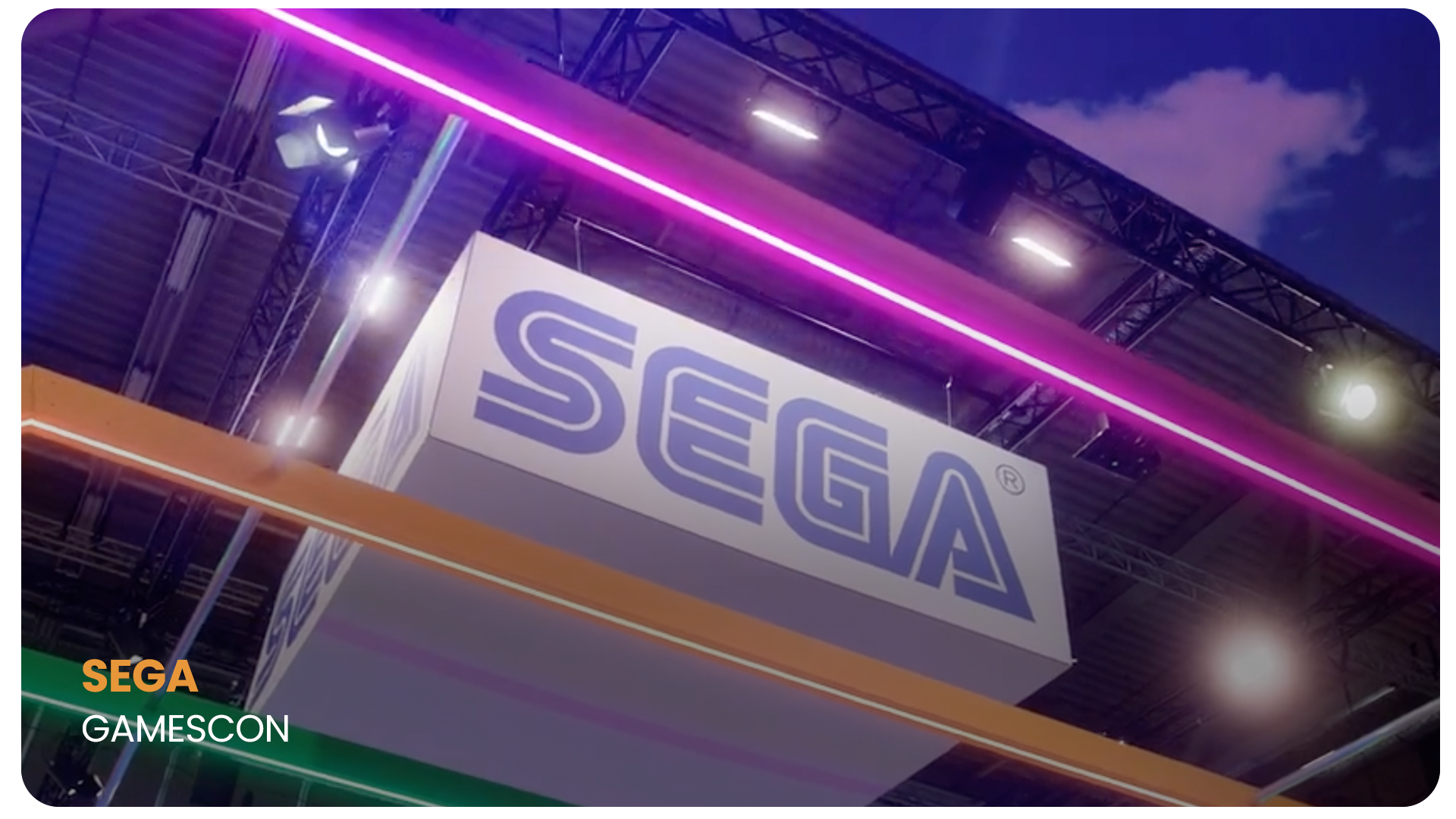 Inside a convention hall with a large SEGA booth, illuminated by neon purple, pink, and green lights, with a visible ceiling structure and promotional banner hanging from above.