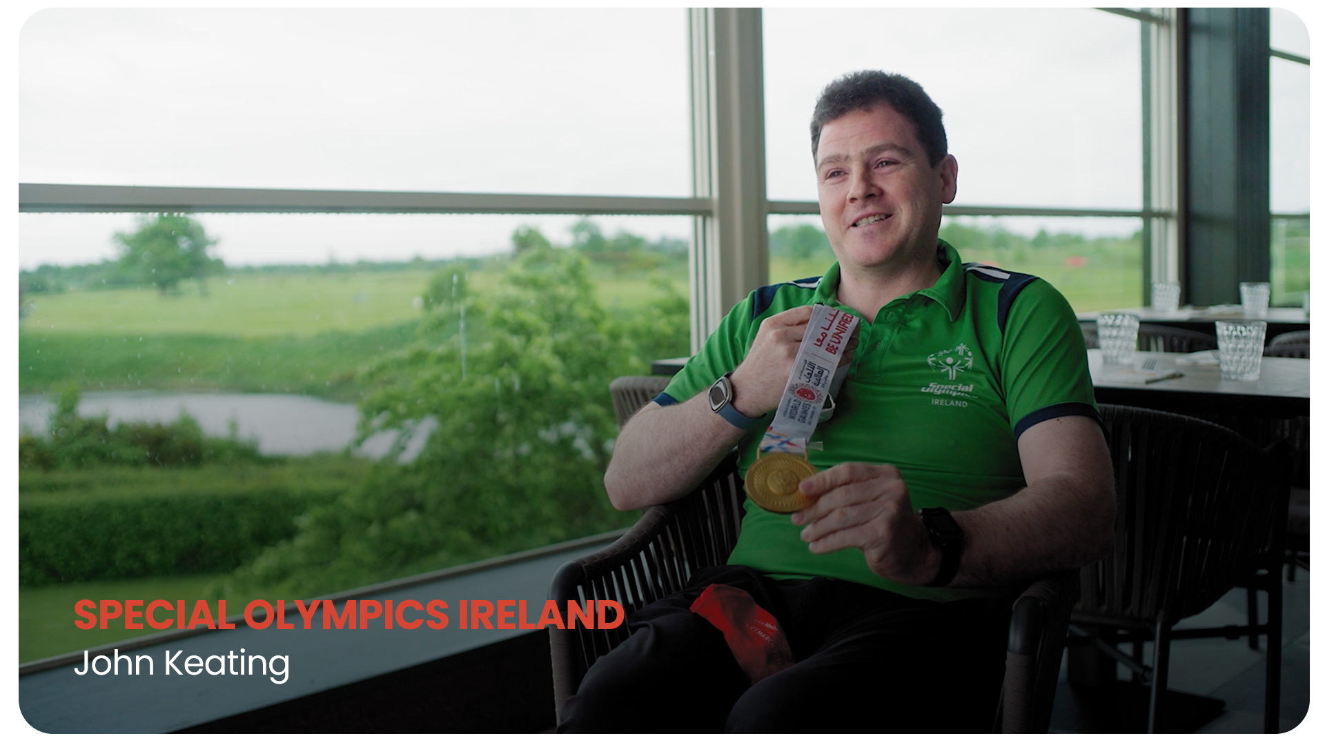 John Keating wearing a green Ireland Special Olympics shirt, holding a gold medal, sitting in a room with large windows overlooking a green landscape, smiling.