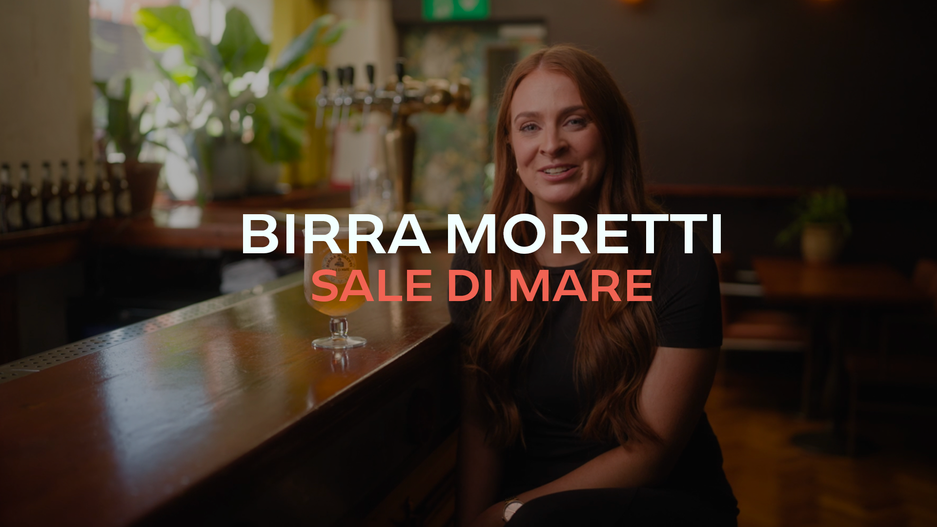 A woman with long red hair sitting at a bar with a glass of beer in front of her. The background has plants and bar equipment. Overlaid text reads, 'Birra Moretti Sale di Mare.'