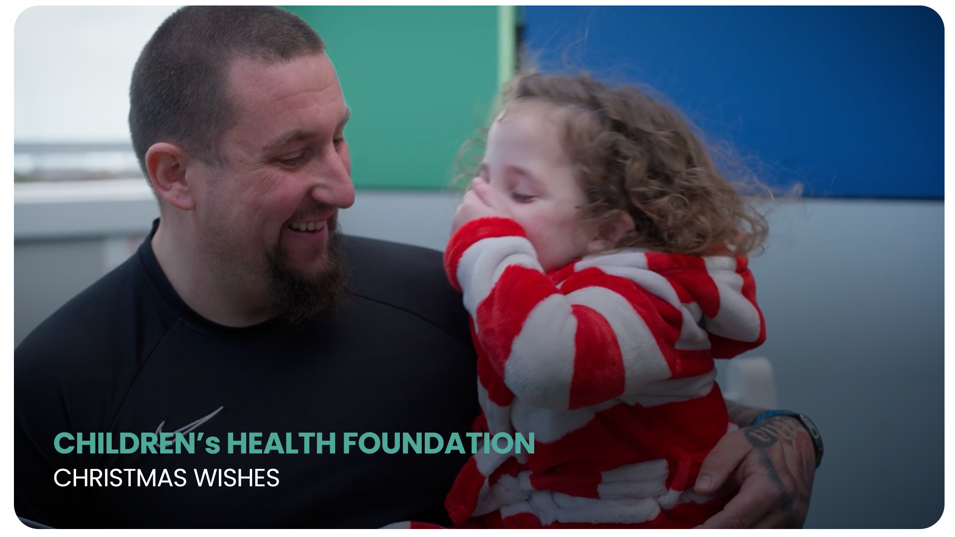 A man with a beard and short hair smiling while holding a young girl with curly hair, dressed in a red and white striped hoodie, who is covering her face with her hand. The background features colorful panels and a window.