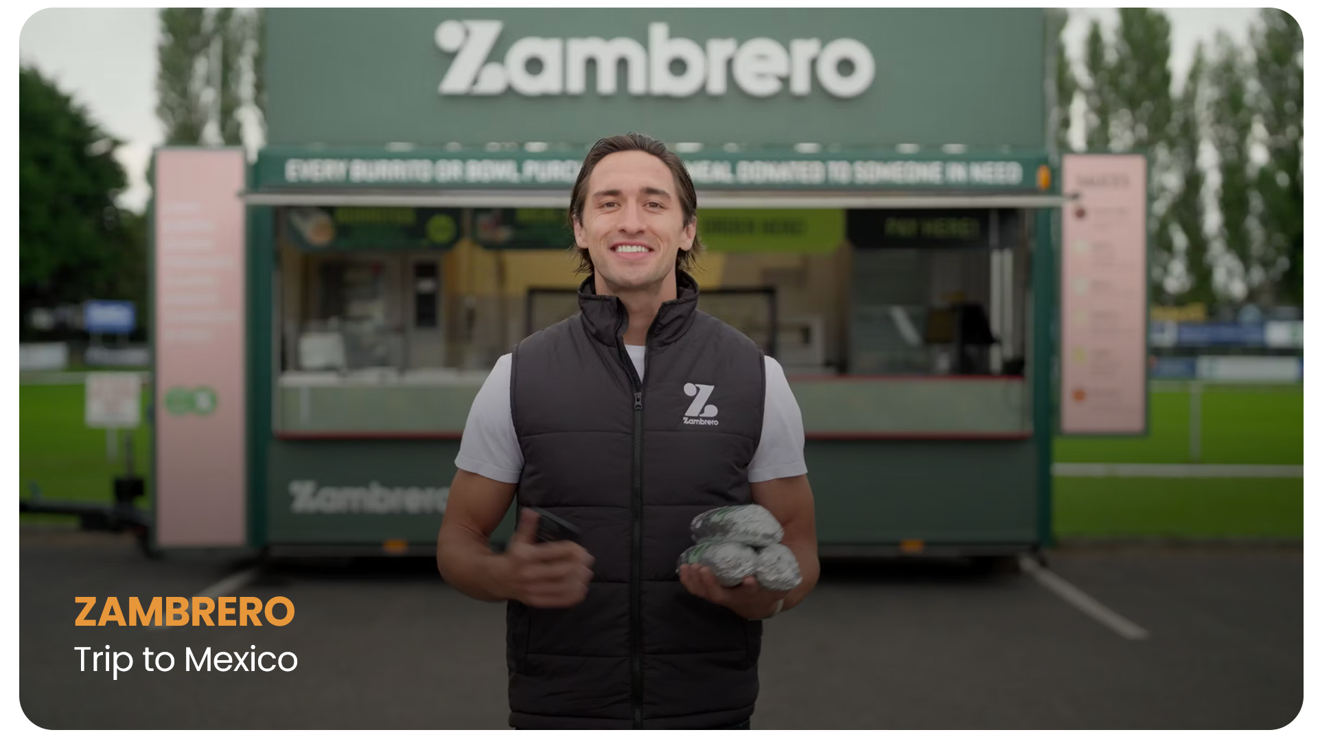 A man smiling at the camera holding a smartphone in one hand and a stack of wrapped food items in the other, standing in front of a green food truck with Zambrero branding, during a trip to Mexico.