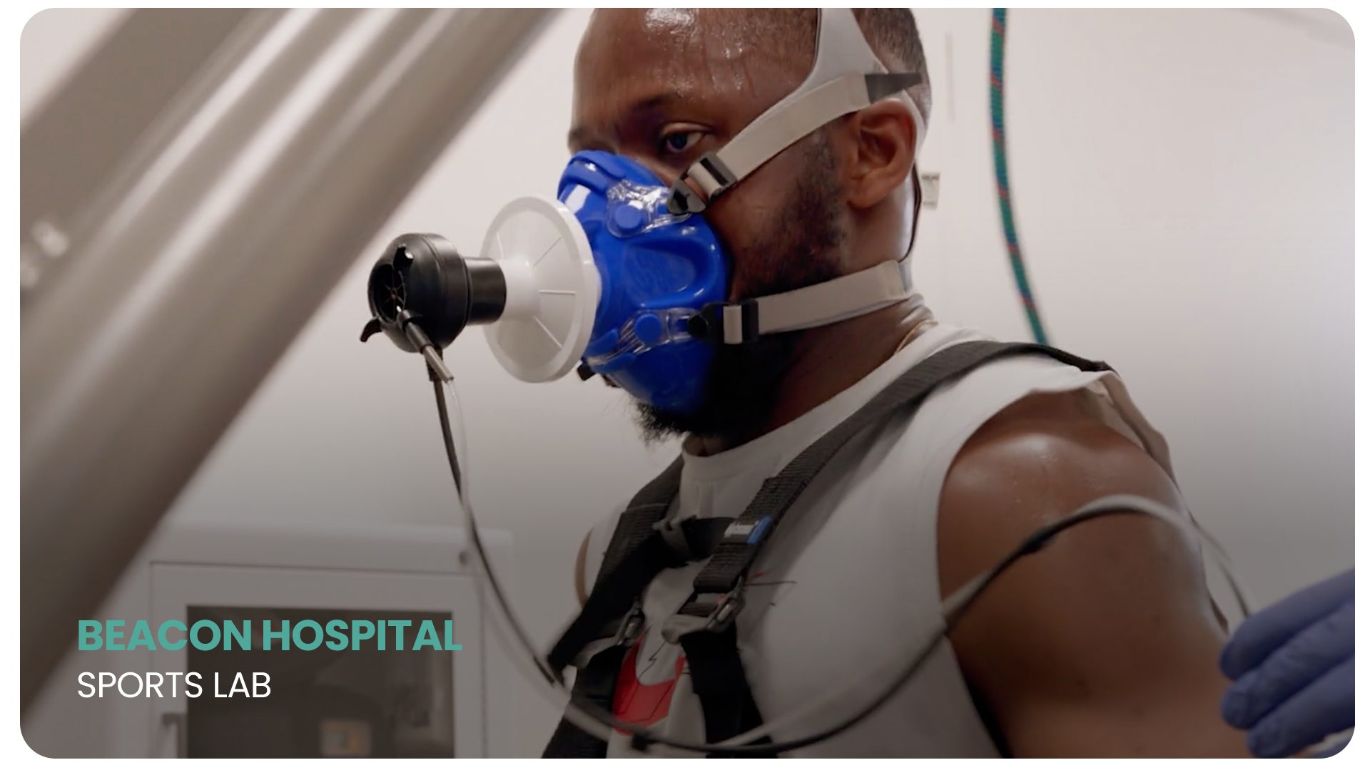 A man with a beard wearing a blue protective mask and a harness, sitting in a sports lab at Beacon Hospital, with a medical or testing device attached to his arm.