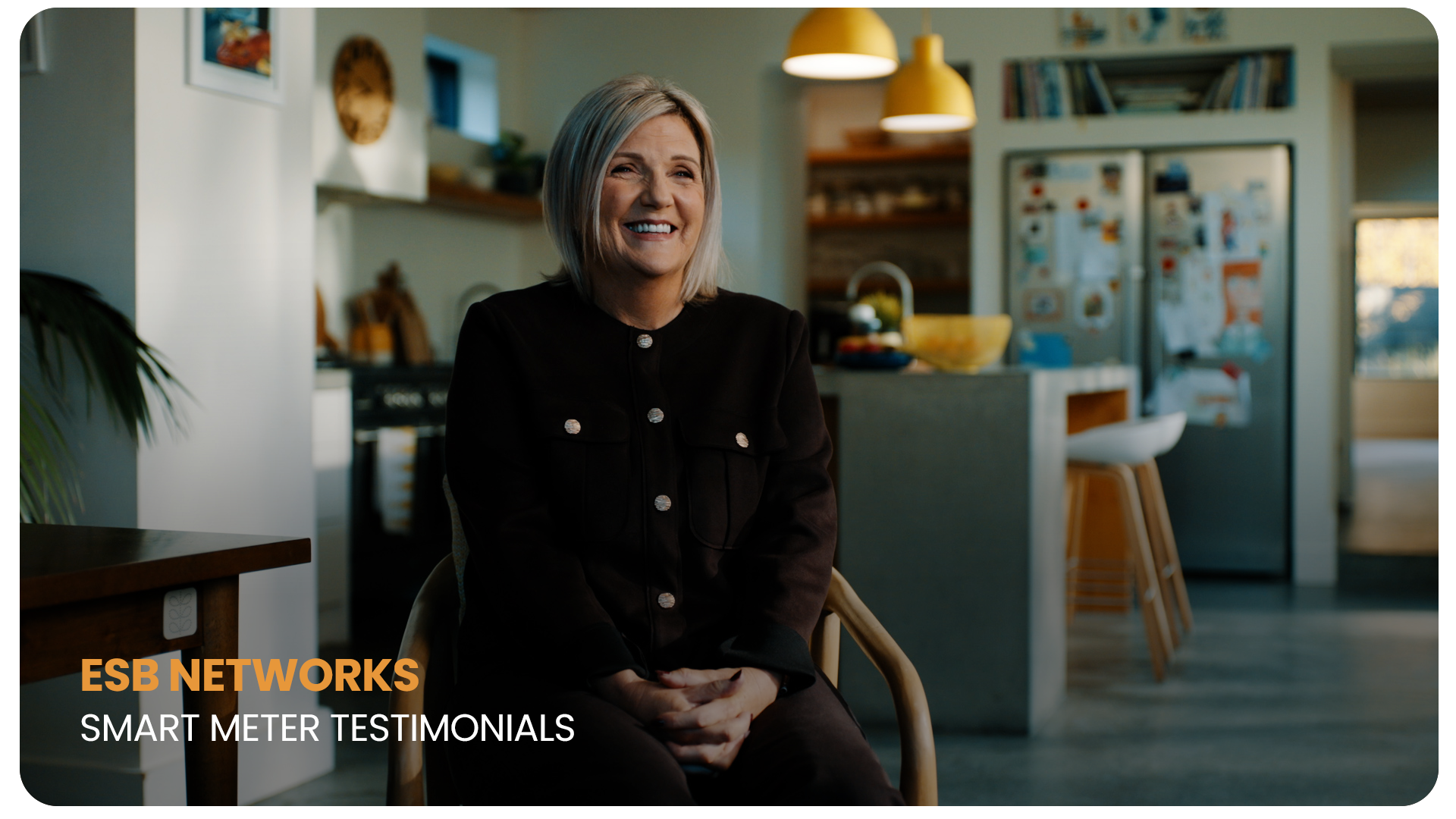 A woman with blonde hair, wearing a black jacket, sitting in a kitchen and smiling during a testimonial interview, with kitchen appliances and decor in the background.