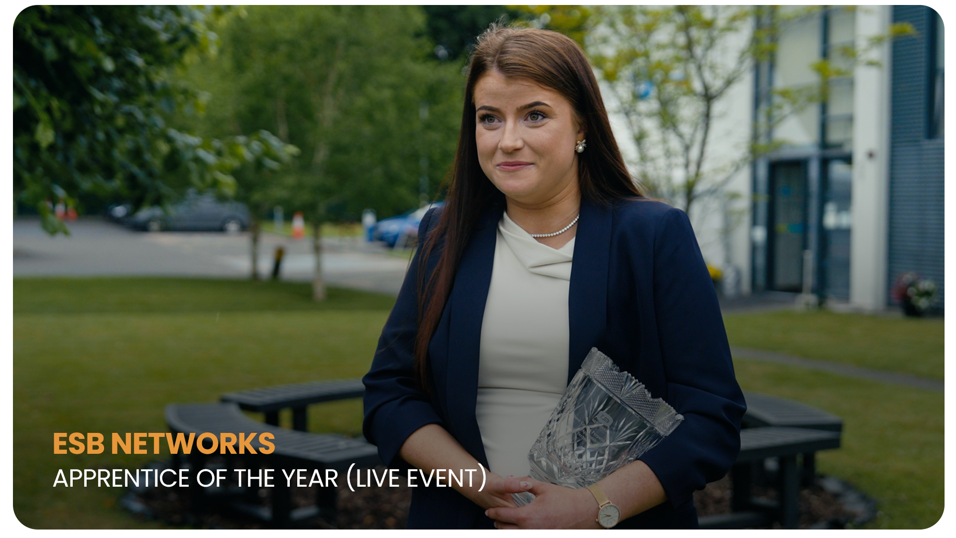 Woman in formal attire holding an award outside, with trees and a modern building in the background, during a professional event.