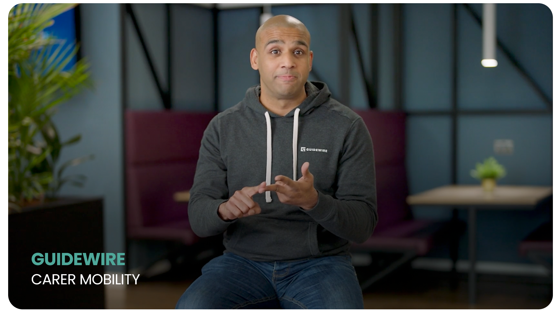 A man sitting in a modern indoor setting wearing a dark gray Guidewire hoodie, making a gesture with his hands, with a plant on the left and a small table with a potted plant in the background. Text on the screen reads "Guidewire Carer Mobility."