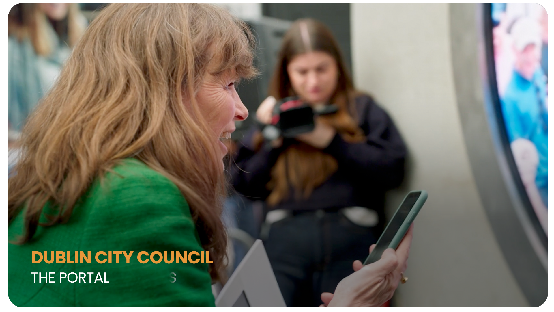 A woman with red hair and a green top is smiling while looking at her phone. In the background, a girl with long brown hair in a black jacket is holding a device, sitting next to a wall and a screen.