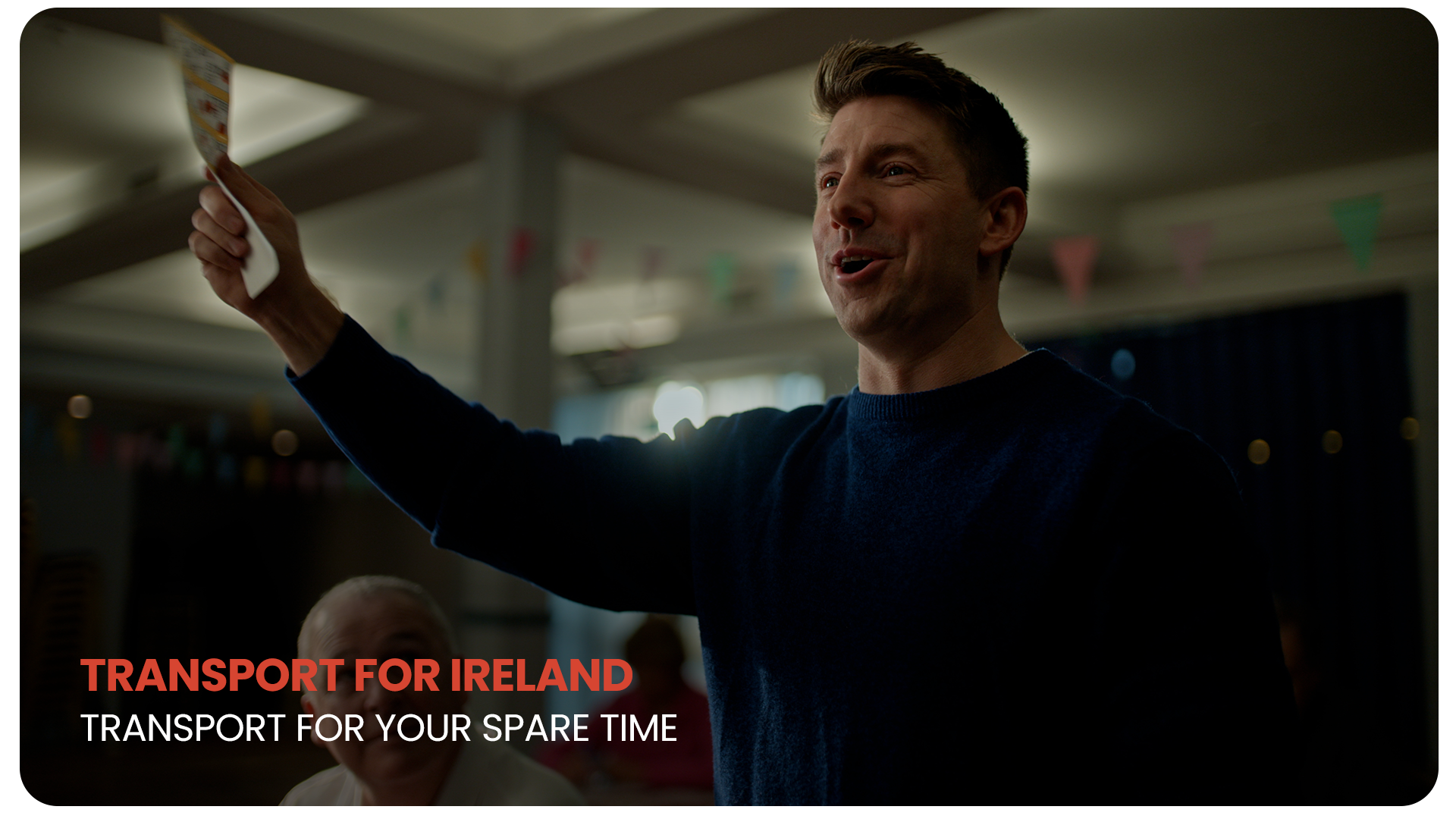 A man speaking at an event, holding up a flyer or card in his right hand, with colorful banners hanging in the background.