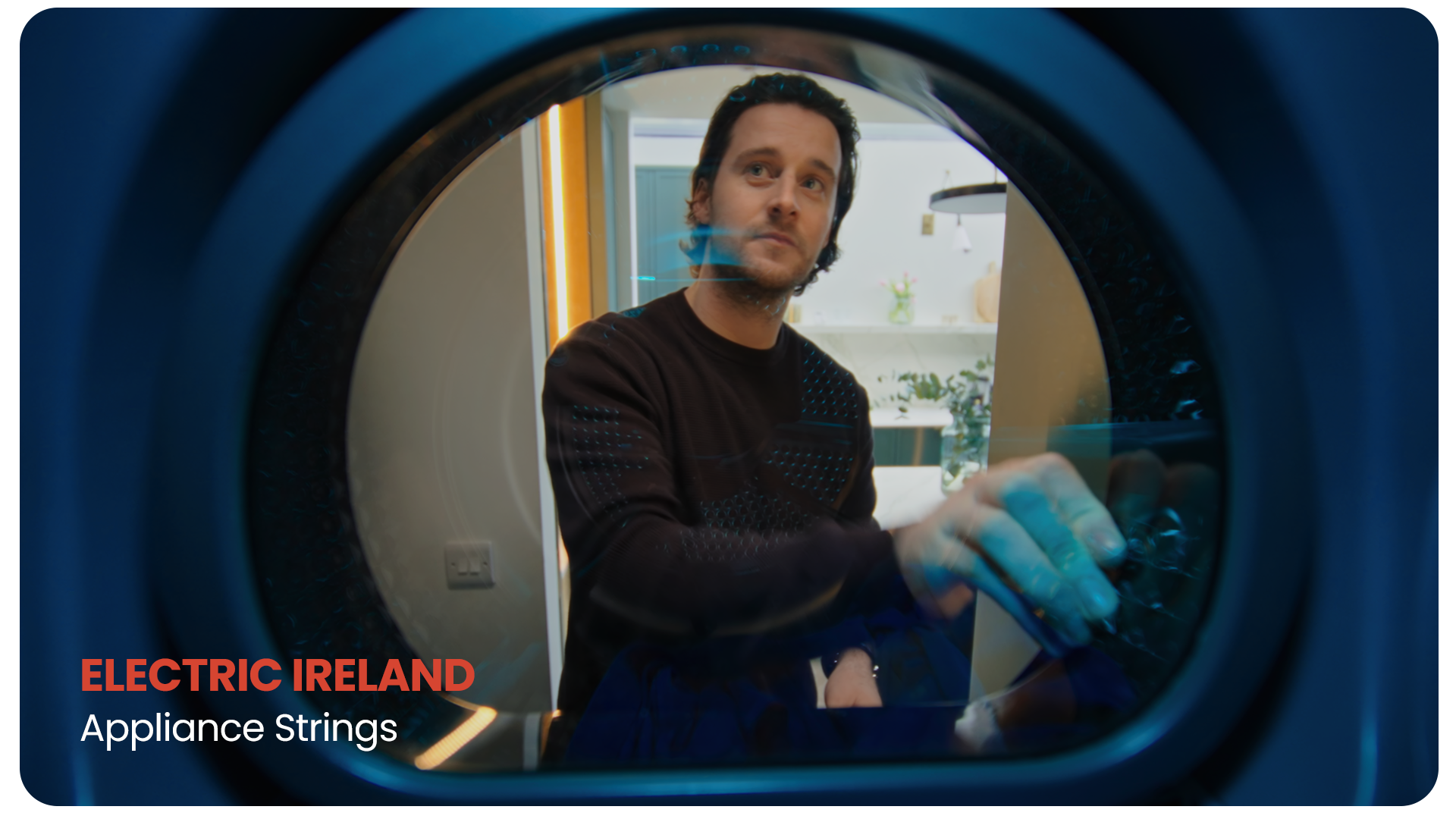 A man is looking into the front of a washing machine, with the door open, as seen through the glass.