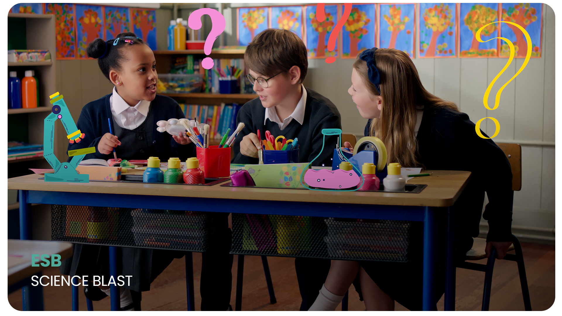 Three young students in school uniforms are seated at a desk in a classroom, engaging in a science activity with colorful craft supplies, plastic bottles, and drawing tools. The classroom background includes colorful artwork on the walls and shelves 
