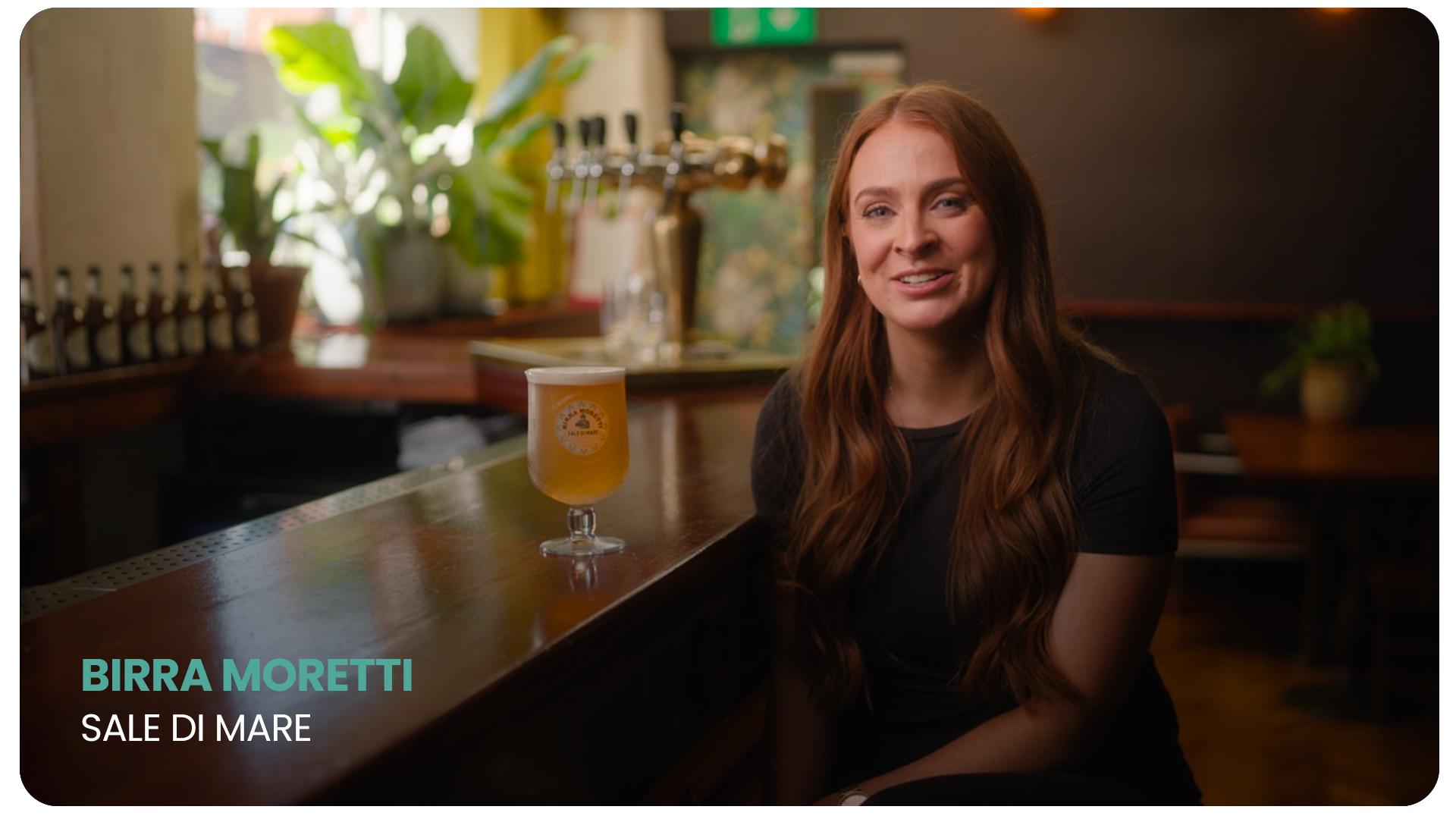 A woman with long red hair sitting at a bar with a glass of beer in front of her, smiling at the camera, in a cozy pub-like setting with plants and beer taps in the background.