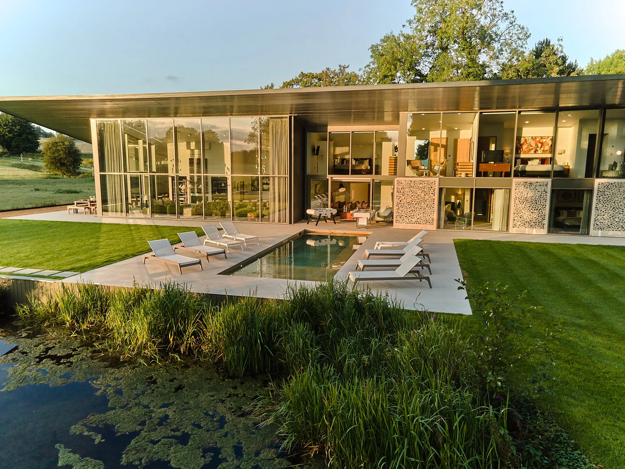 Stillwater's House with large glass windows overlooking a pond and green lawn, with trees in the background, during sunset.