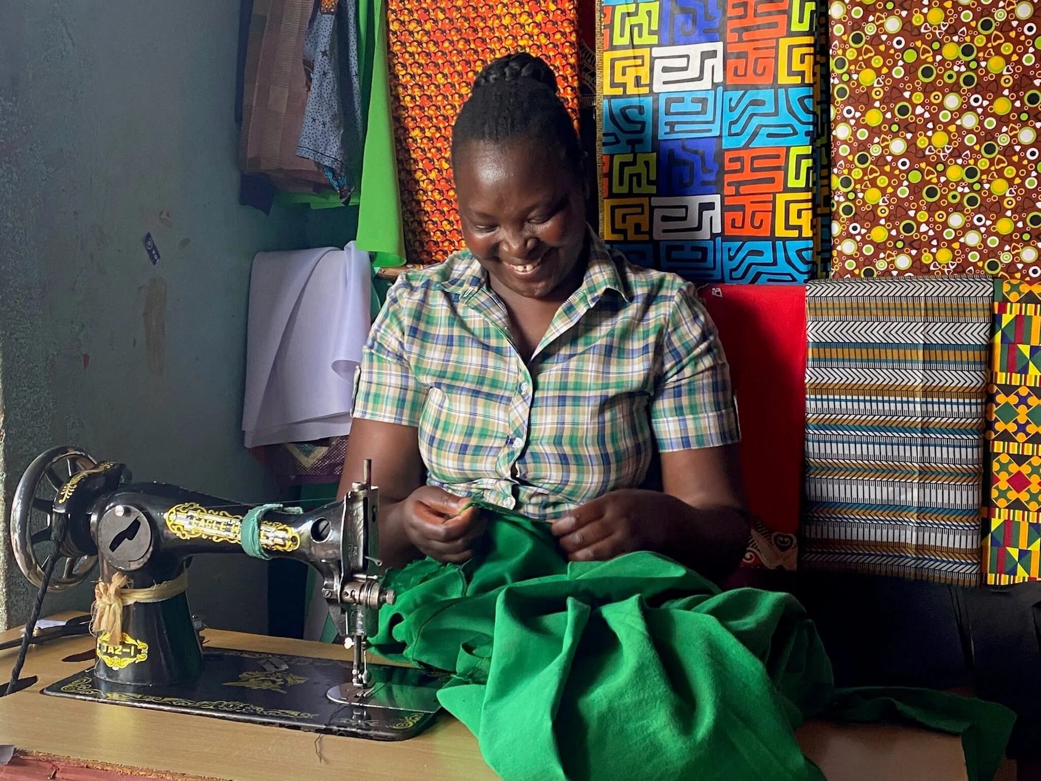 A person wearing a plaid shirt, is sitting at a sewing machine sewing bright green fabric. The background showcases vibrant, colorful African textile patterns and fabrics on display. Recipient of root start up program by The Boo Charity 
