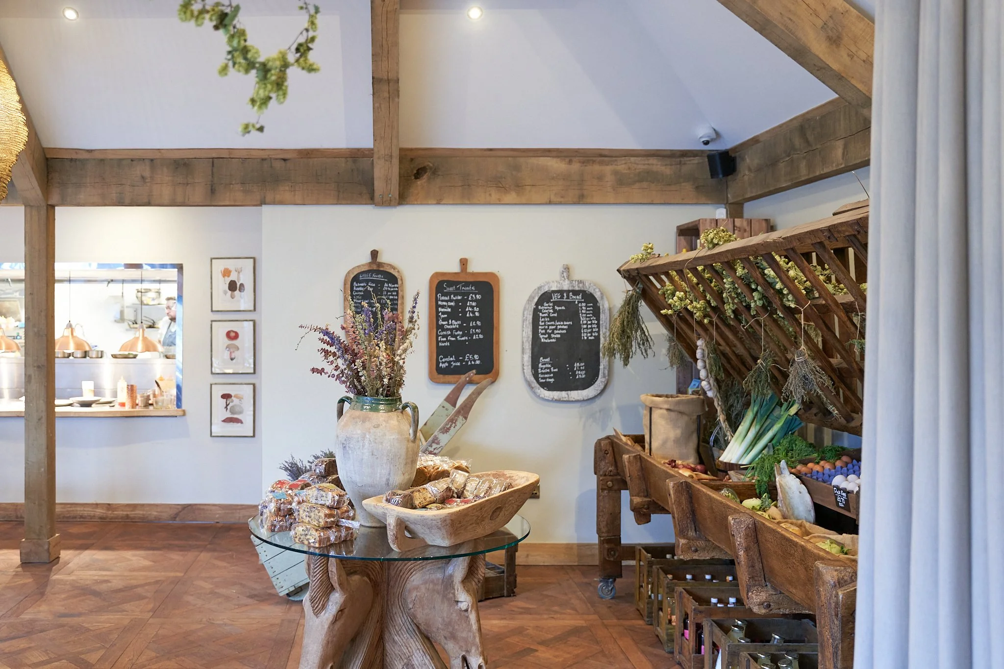 Interior of The Chestnut Horse shop with wooden beams, chalkboard menus, a pottery vase with dried flowers on a glass table, and wooden shelves displaying fresh produce and eggs.