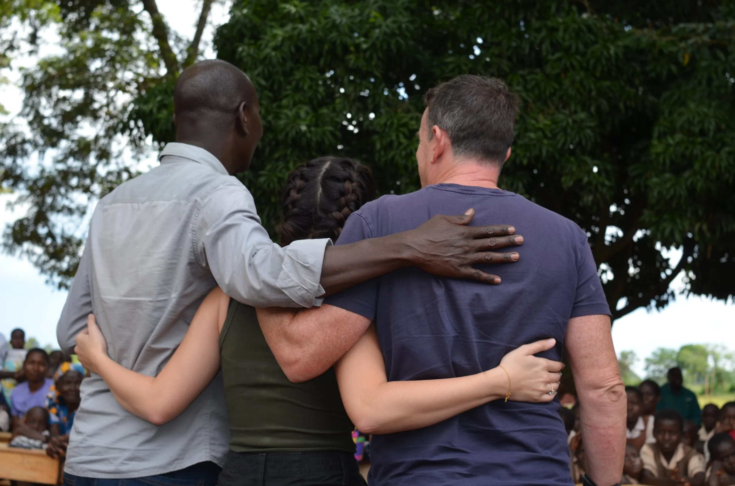 People standing close together in an outdoor setting, with their arms around each other, showing unity and support. In the background, a group of onlookers watches. The Boo Charity