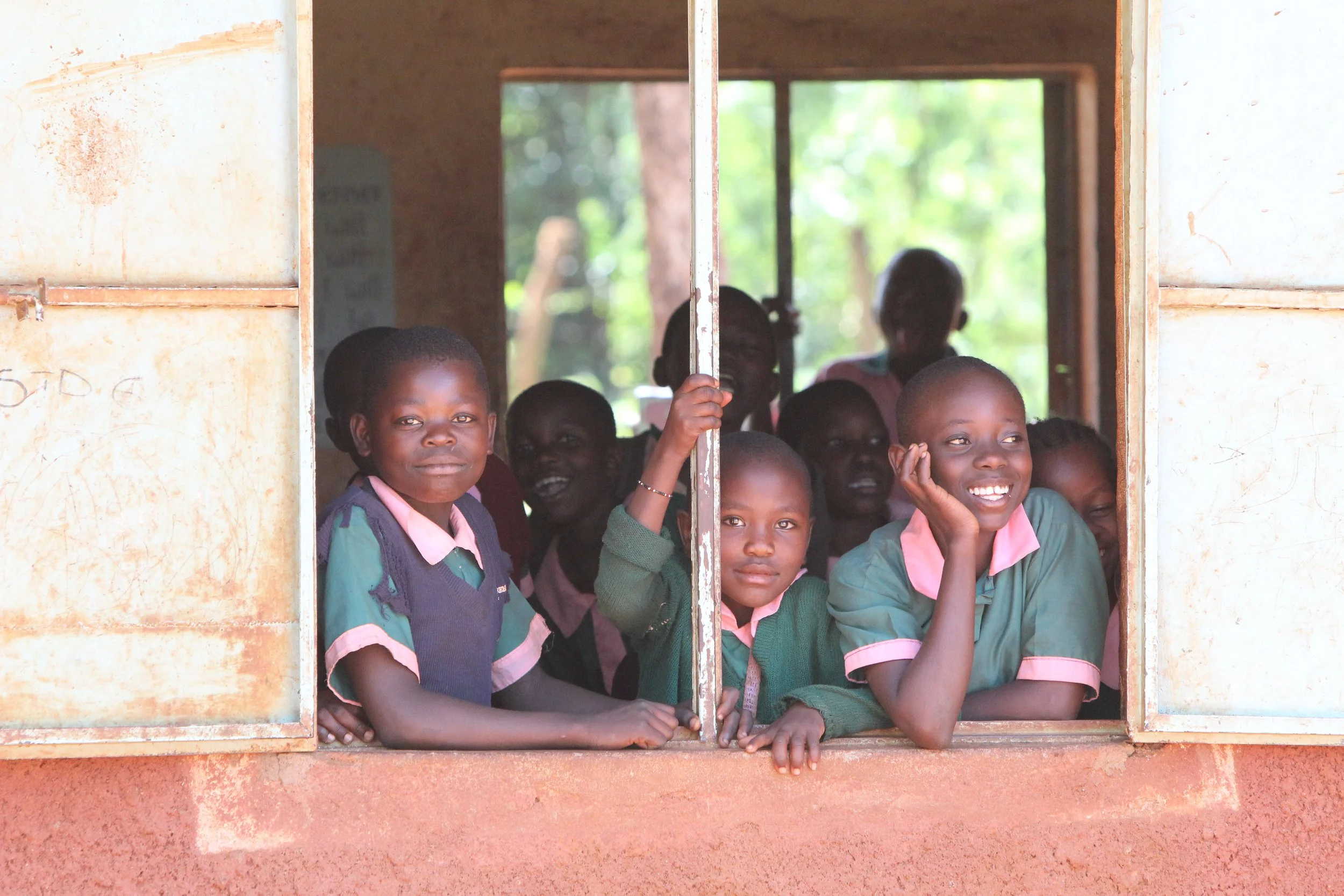 Group of children at a project that The Boo Charity sponsors, looking out from a classroom window with a rural outdoor background visible through the window.