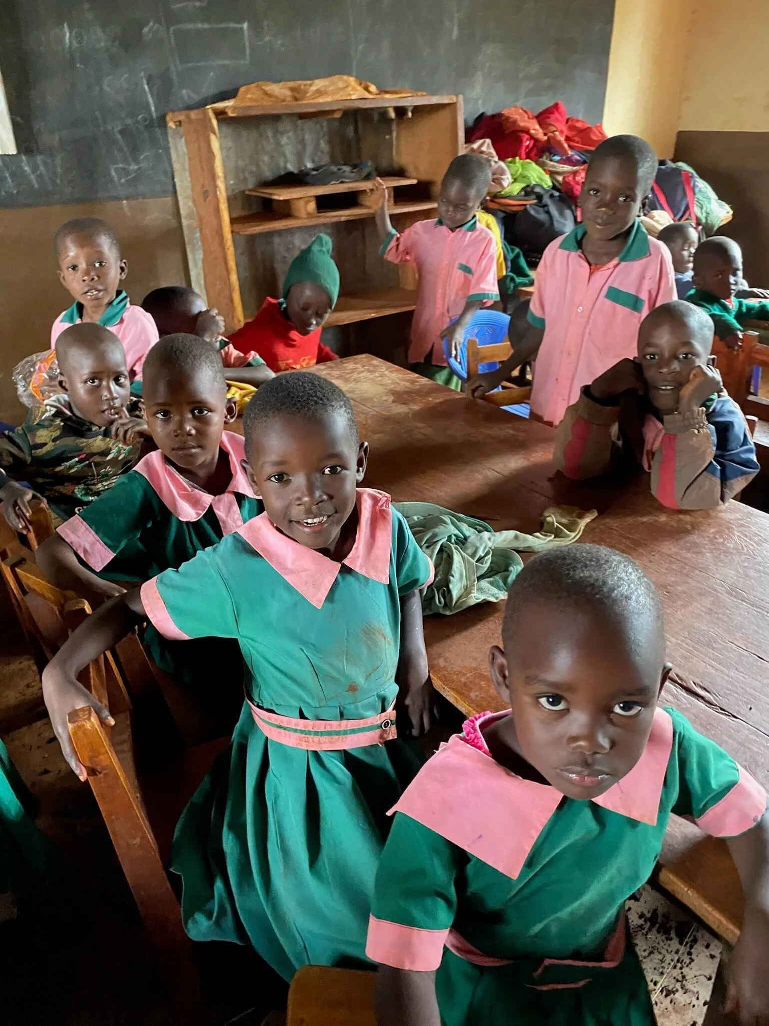 Group of young children in colorful school uniforms inside a classroom, sitting and standing around wooden tables with a chalkboard on the wall. Project sponsored by The Boo Charity.