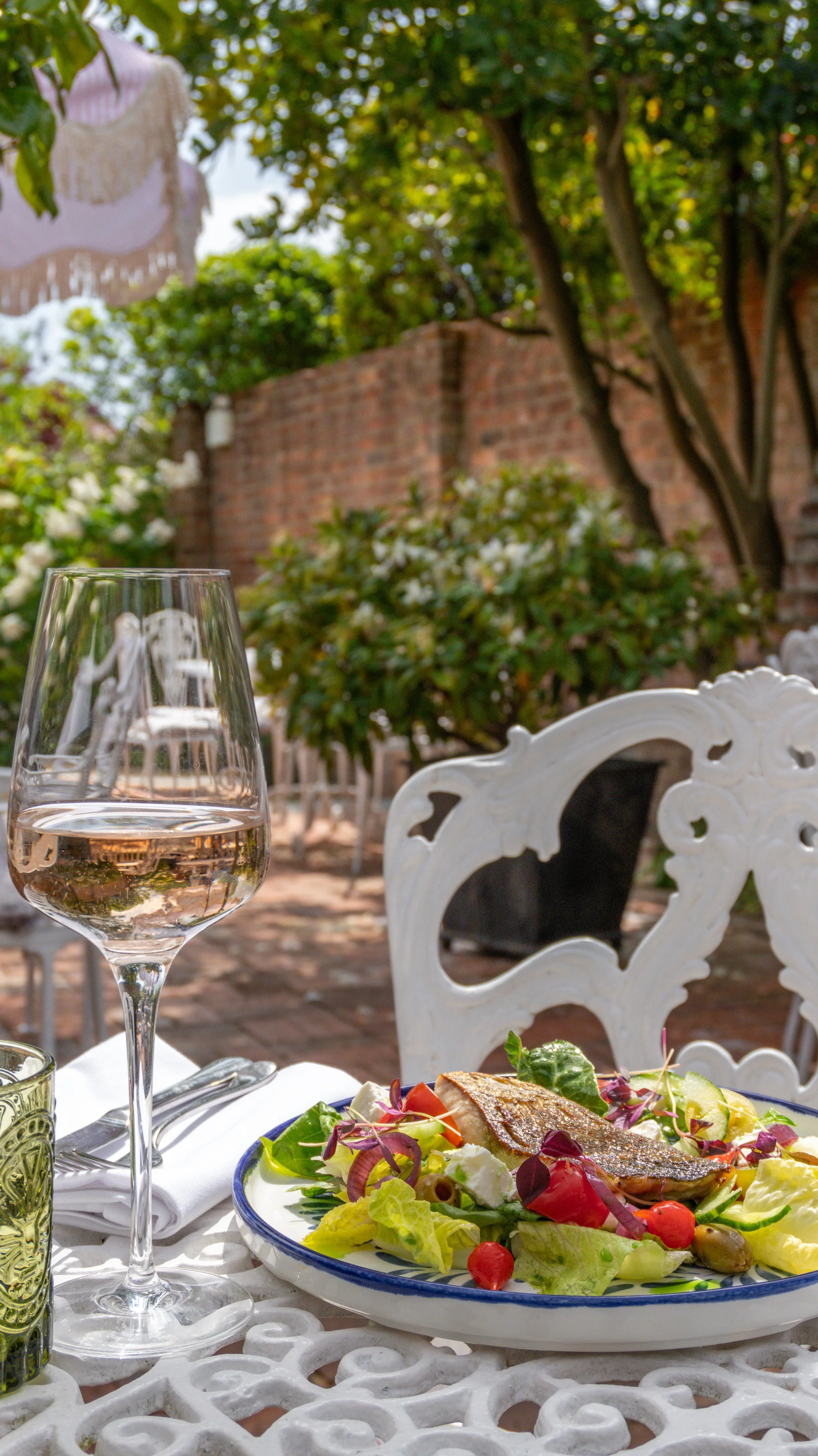 A glass of rose, a plate of salad with grilled fish, and a glass of water on a white outdoor table in The Garden at Stanwell House