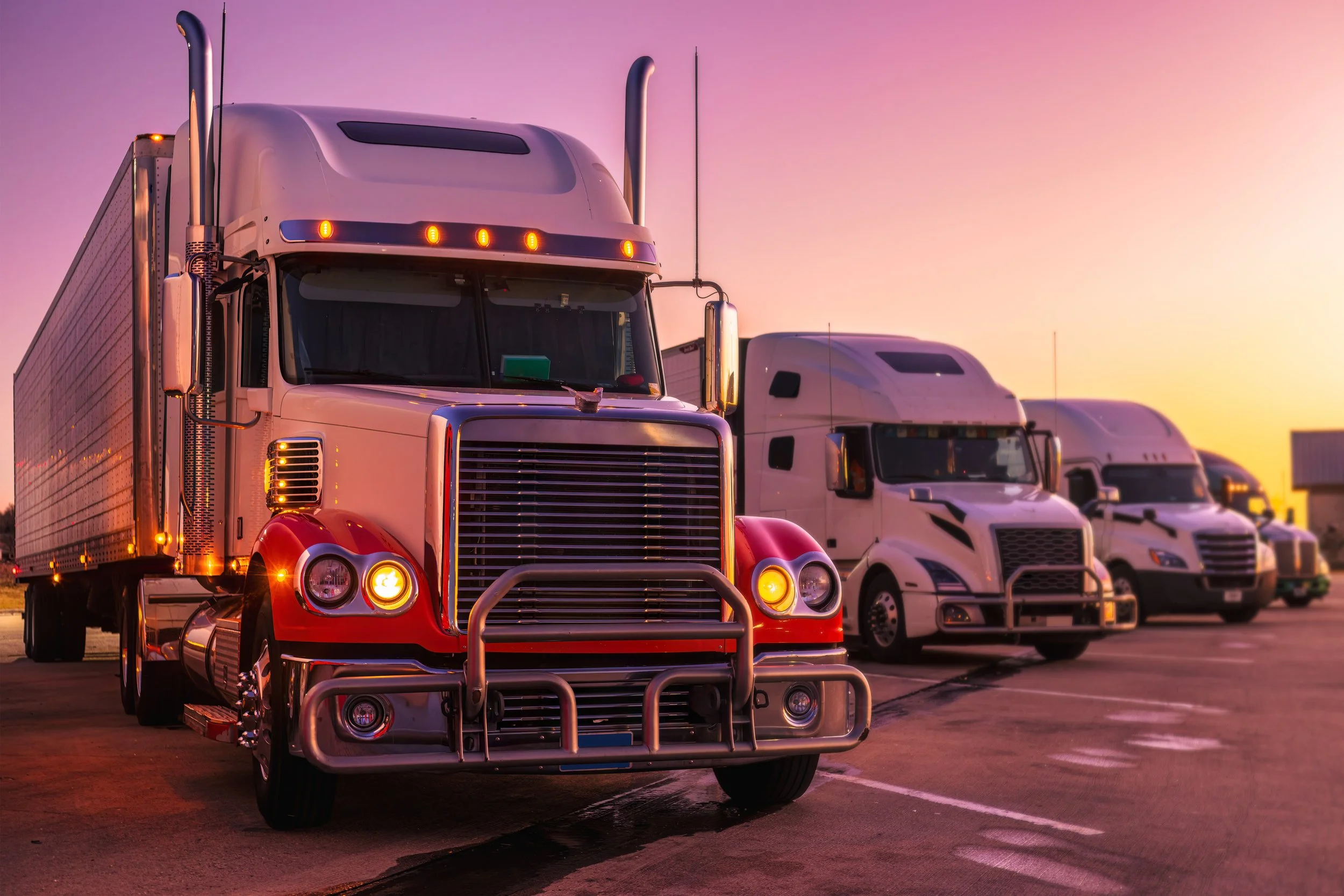 A row of semi-trucks parked in a lot at sunset, with colorful sky in the background.