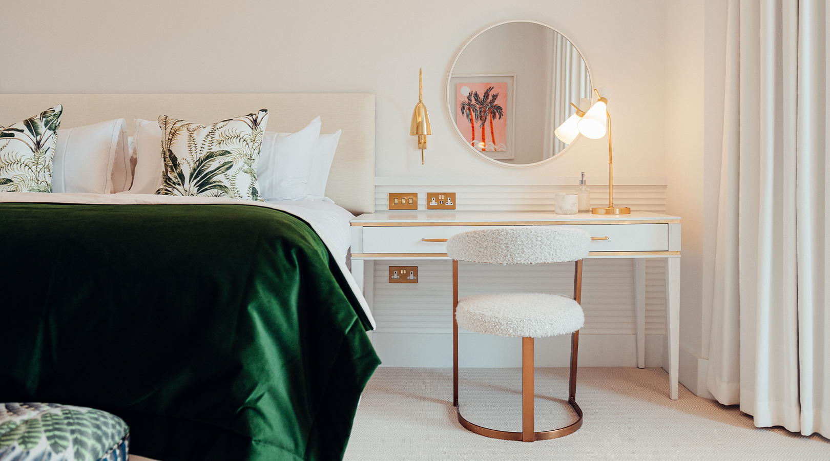 Bedroom at THE NICI with a white desk, round mirror, plush chair, and bed with tropical print pillows, adorned with gold accents