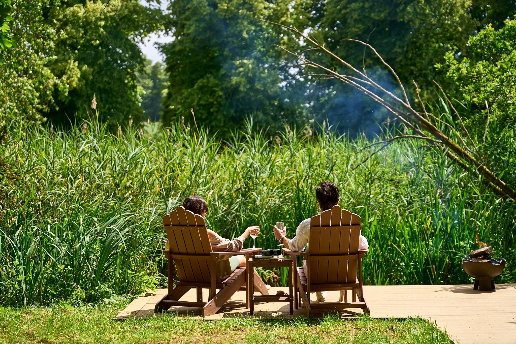 A couple sitting on wooden chairs on a deck at Avington Estate, relaxing with glasses of wine, facing a lush green landscape with tall grass, trees, and smoke rising in the distance.