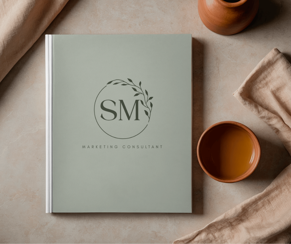 A light green book titled 'Sociabella Media Marketing Consultant' with a logo of a circular branch and leaves, placed on a beige surface. Two terracotta cups and beige cloths surround the book.