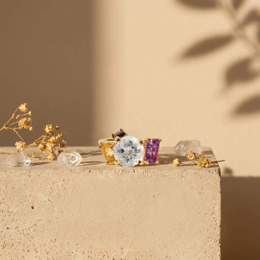 Close-up of Amy Russell Taylor jewelry pieces, including a ring with a large clear gemstone and a ring with a purple stone, placed on a beige surface with dried flowers and small clear stones. Shadow of a plant is visible on the background wall.