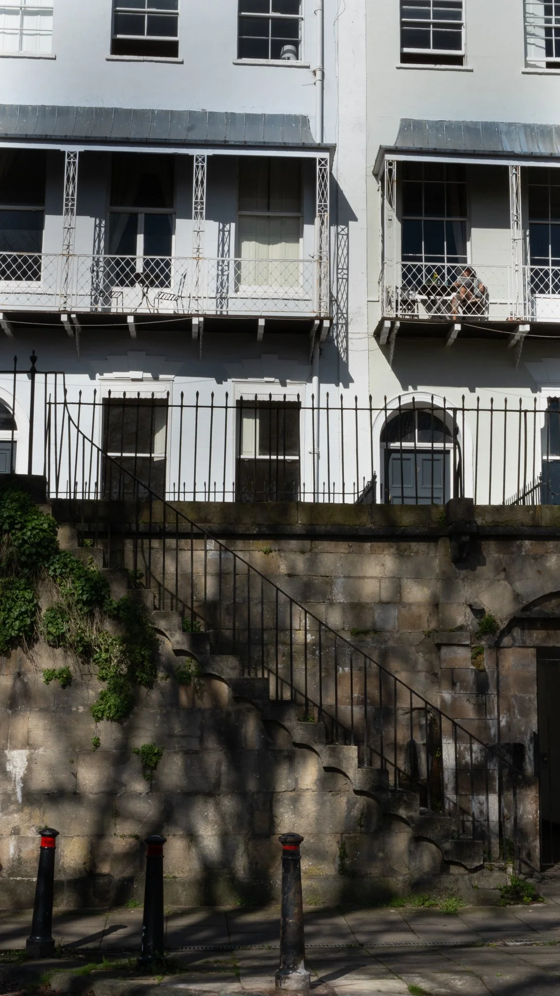 Front view of a multi-story building with balconies, a wrought iron fence, and stairs leading up to the entrance, with shadows cast on the wall. Royal York Crescent, Bristol