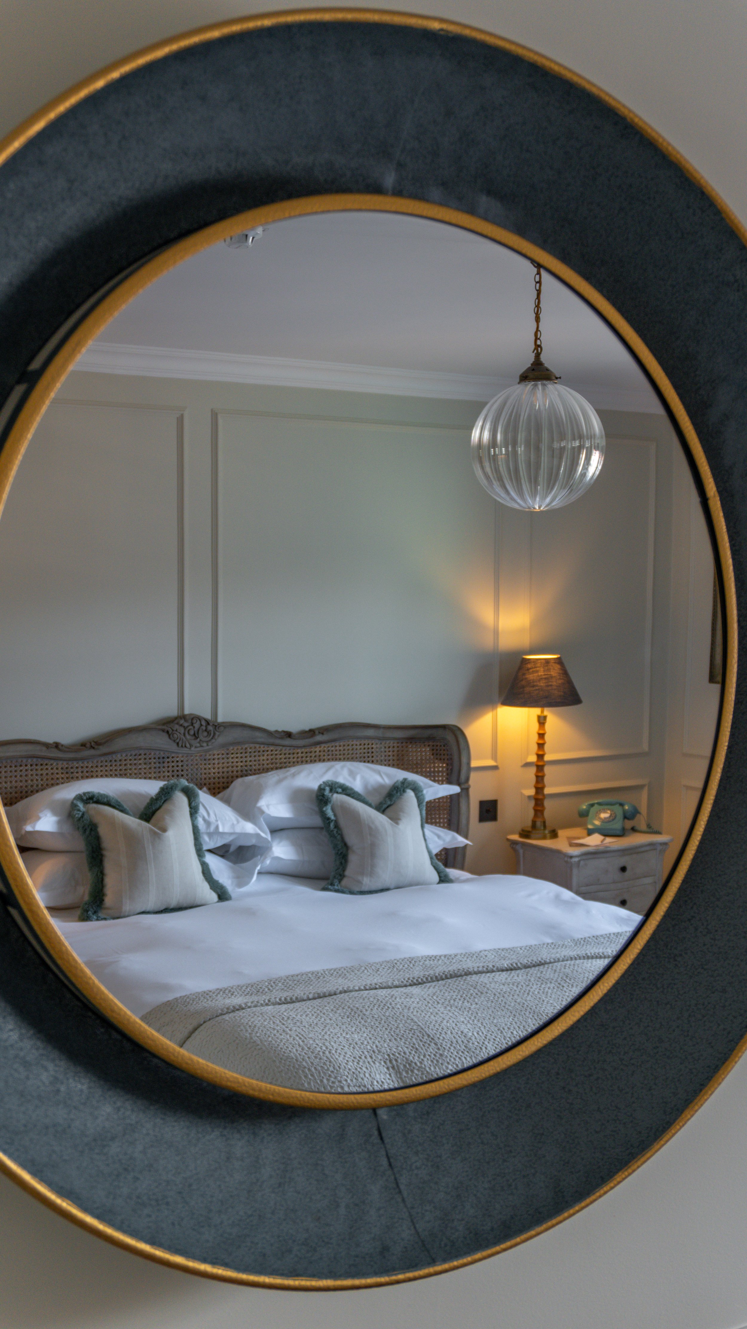 A mirror reflecting a cozy bedroom at Stanwell House with a bed that has white linens and decorative pillows, a nightstand with a vintage telephone, a lamp, and a hanging glass pendant light.