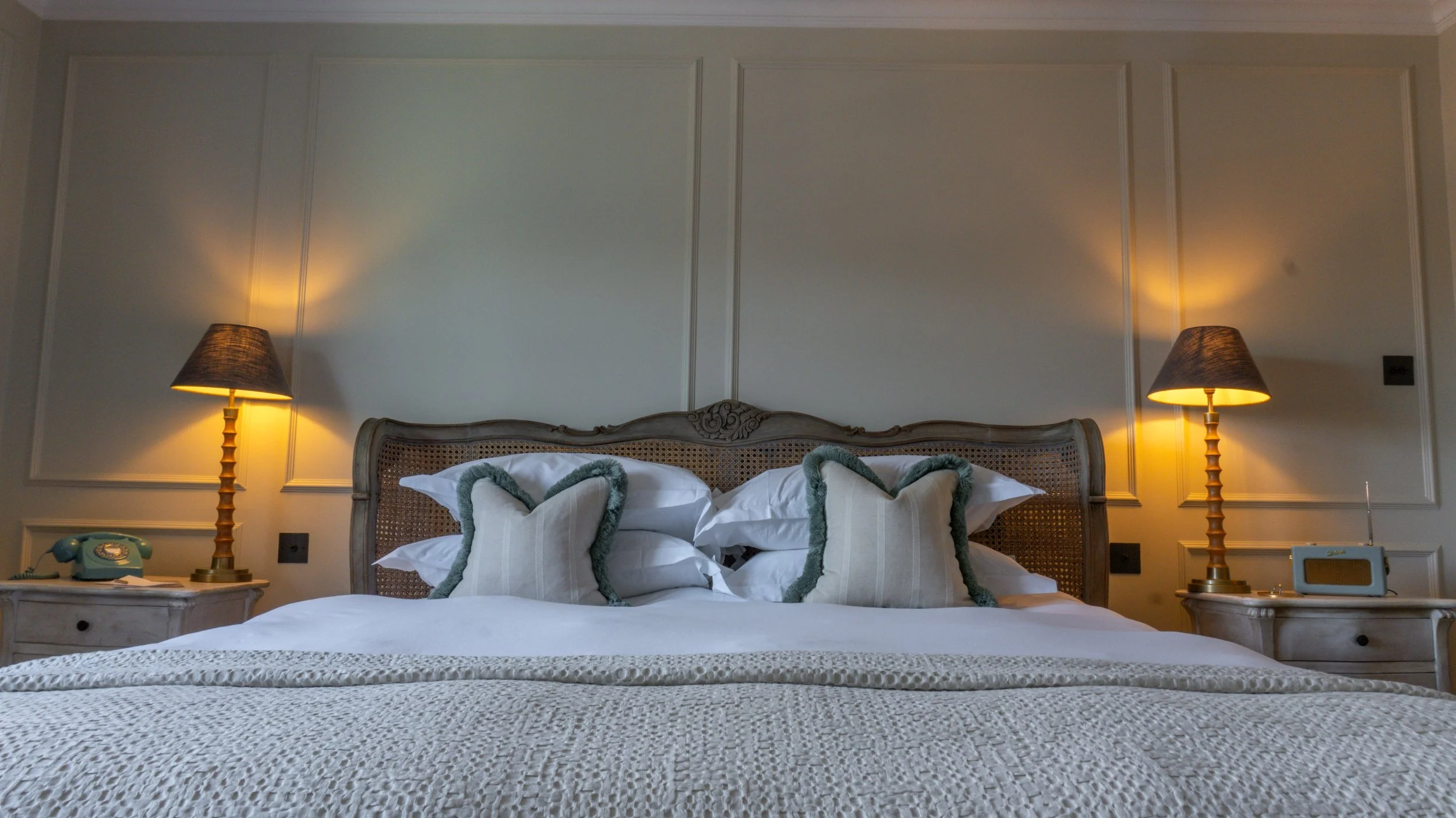 A cozy bedroom at Stanwell House with a large bed that has white bedding and decorative pillows. There are two matching bedside tables with vintage-style lamps and a mint-green rotary phone on the left table.
