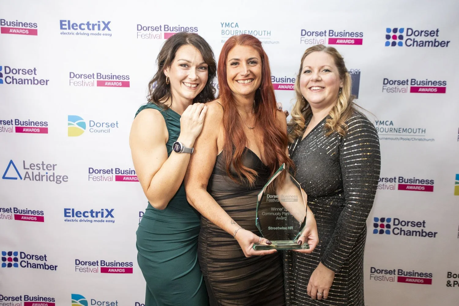 Streetwise HR Gurus at an awards ceremony holding a glass award, smiling in front of a backdrop with various sponsor logos.