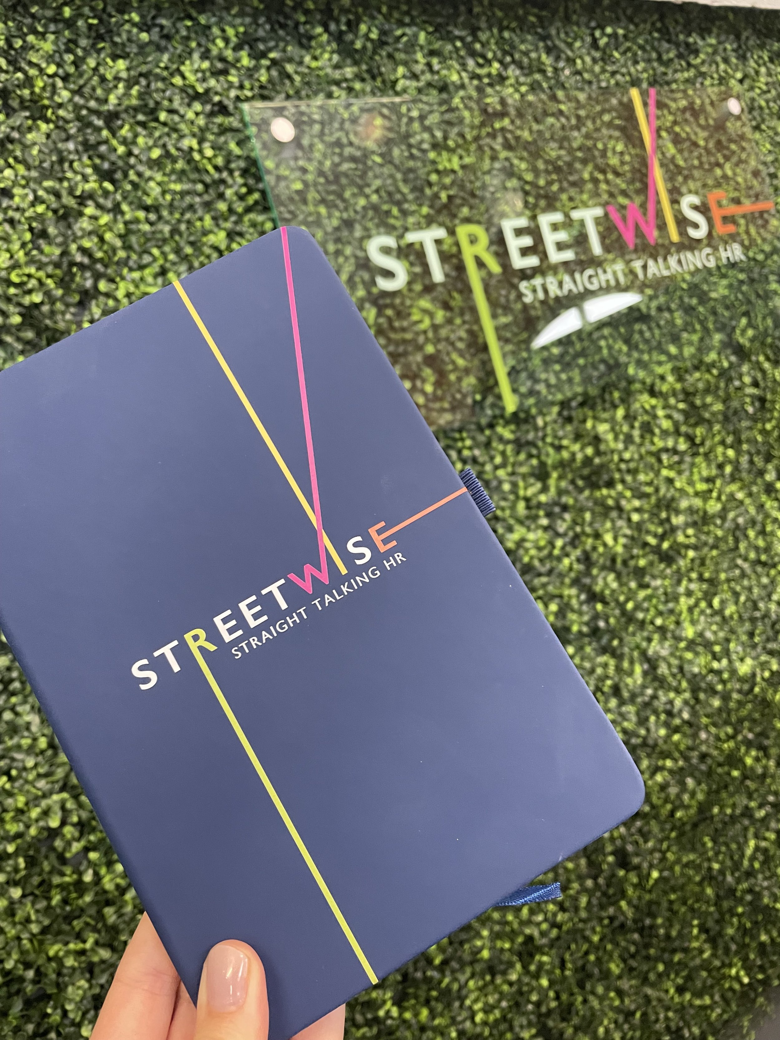 Close-up of a hand holding a navy blue booklet with colorful lines and the text 'STREETWISE STRAIGHT TALKING HR' on the cover, with a glass sign with the same text in the background on a green grassy surface.