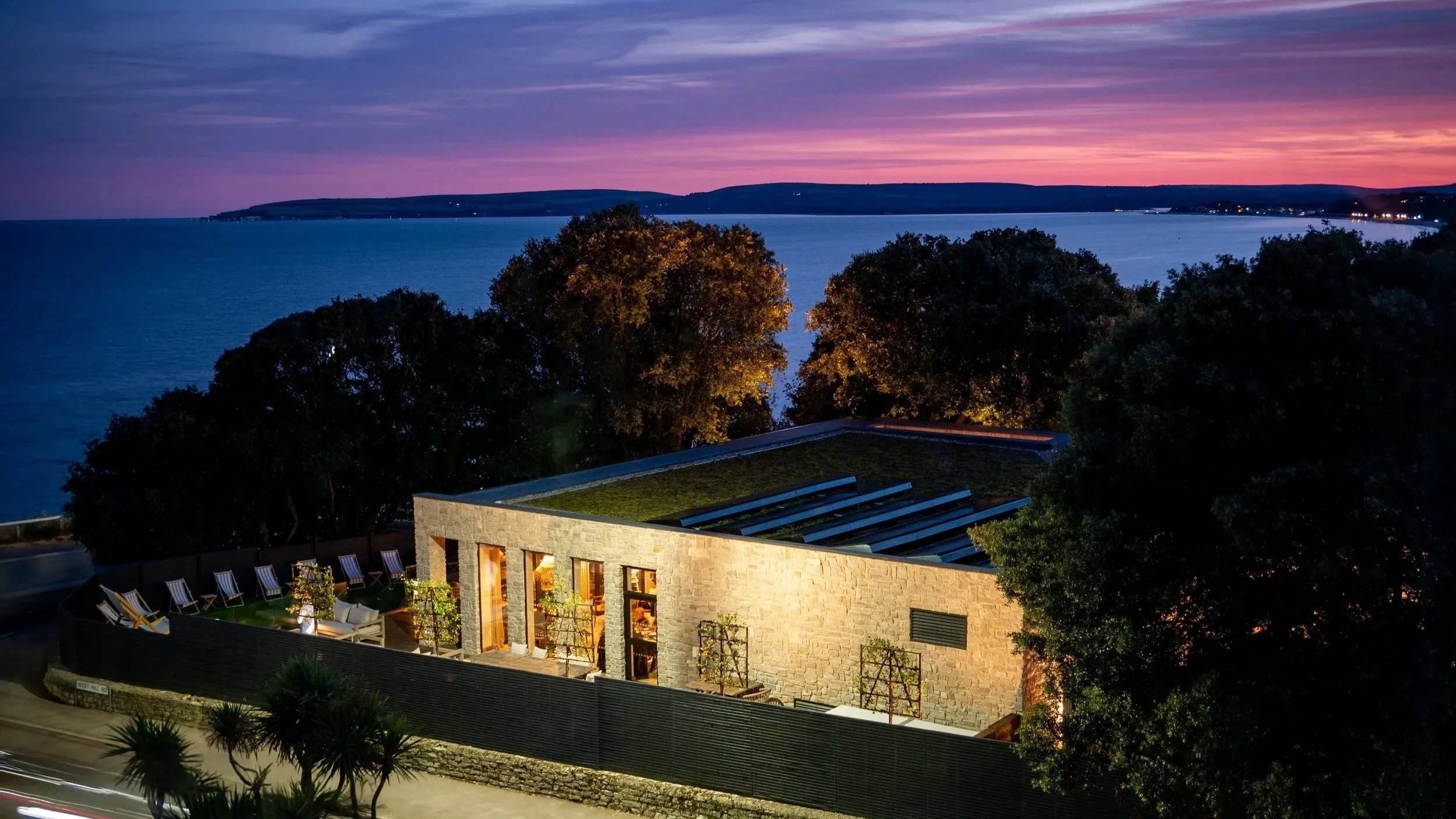 Old Harry Rocks restaurant with large windows and a rooftop garden, overlooking a bay during twilight with colorful sky and distant hills.