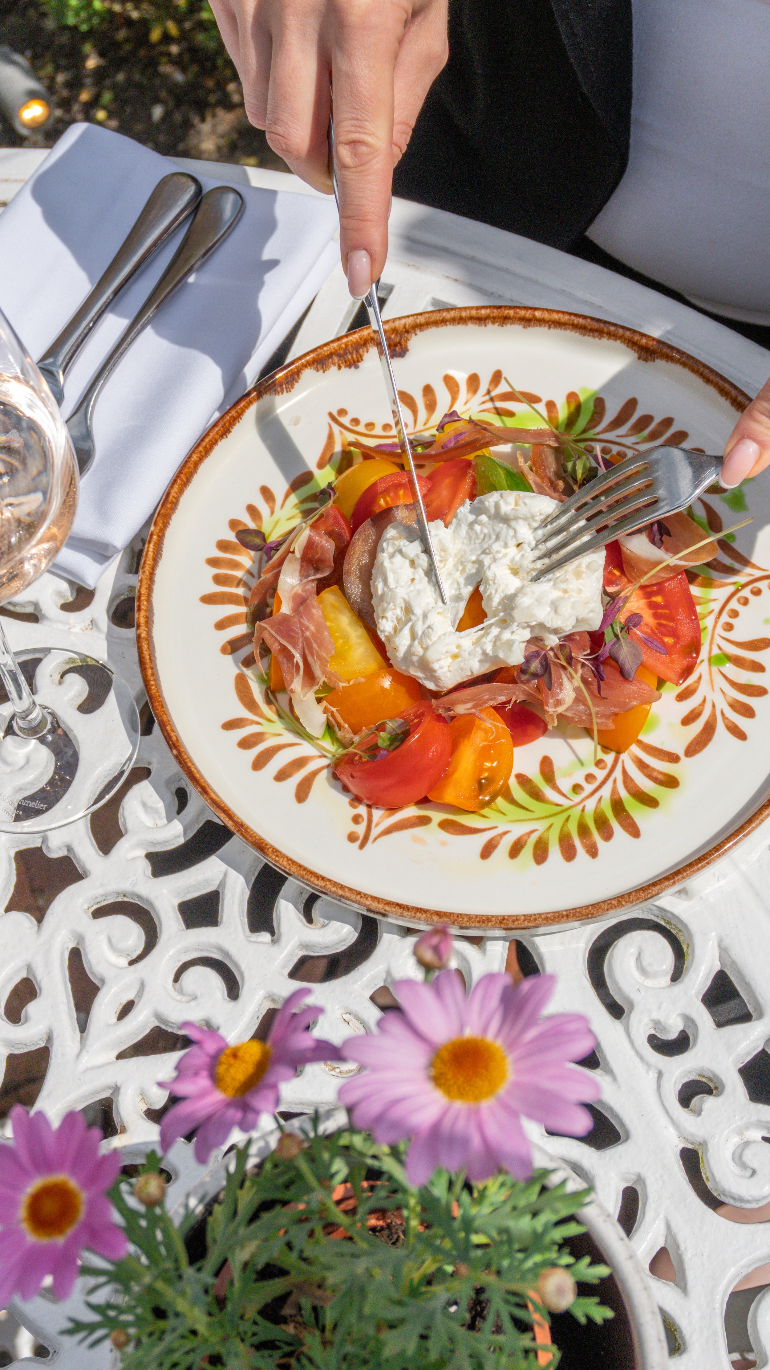 A person is cutting into a colourful salad with a fork and knife. The salad contains burrata tomatoes, prosciutto, and fresh greens. Garden menu at Stanwell House