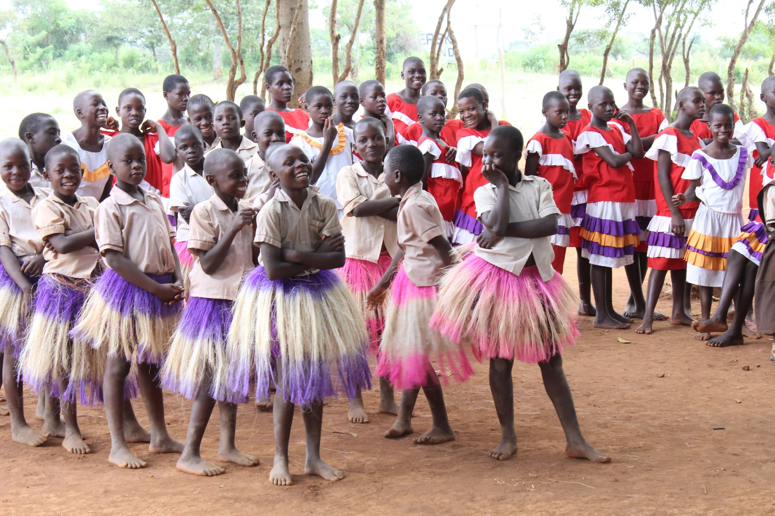 Group of children dressed in colorful clothing, standing outdoors on a dirt ground, with trees in the background. Project sponsored by The Boo Charity.