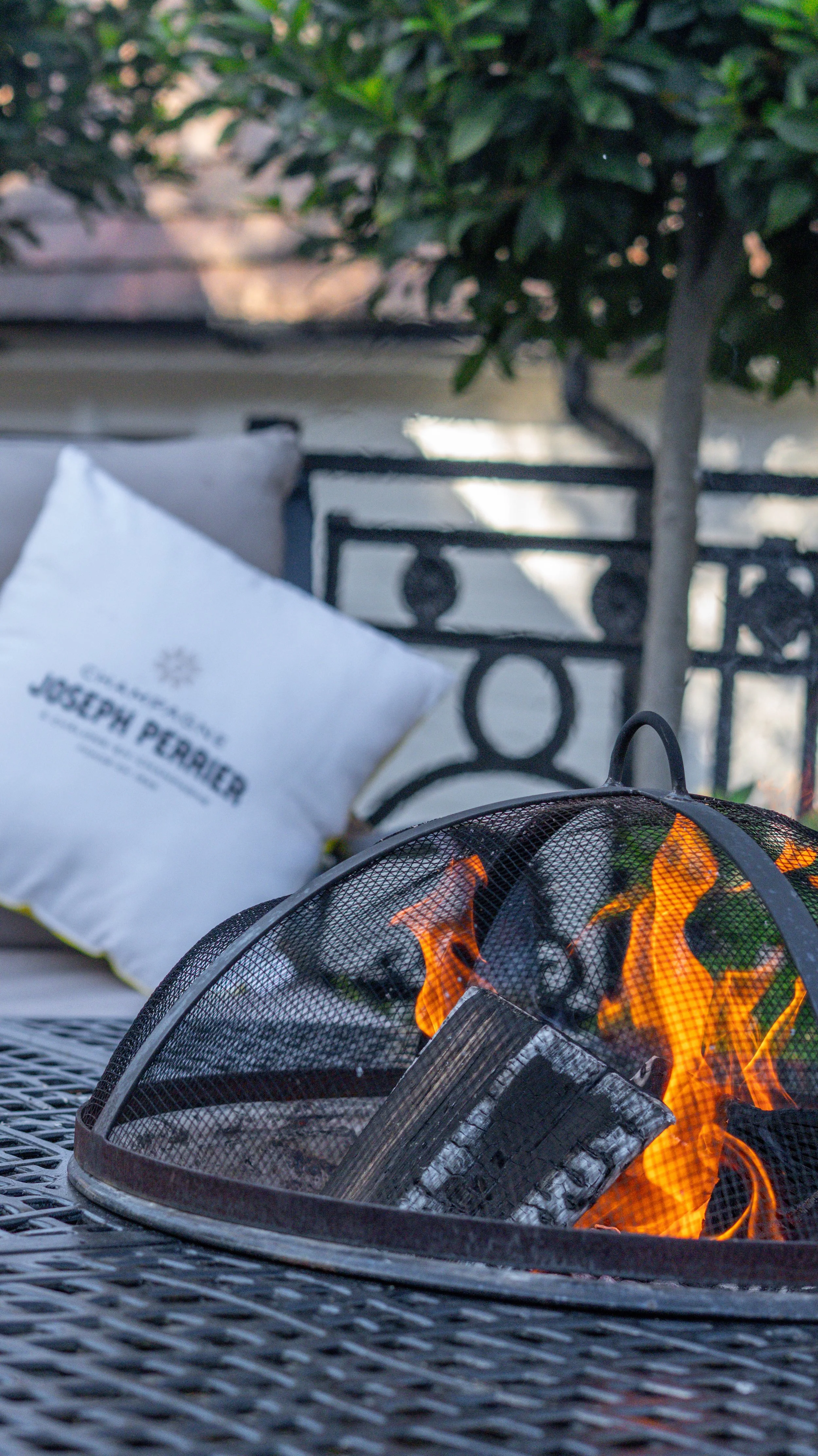 Stanwell House patio scene with a lit tabletop fire pit, a white pillow labeled 'Joseph Perrier' on a seat, a metal table, and a background with lush green bushes and a black metal railing.