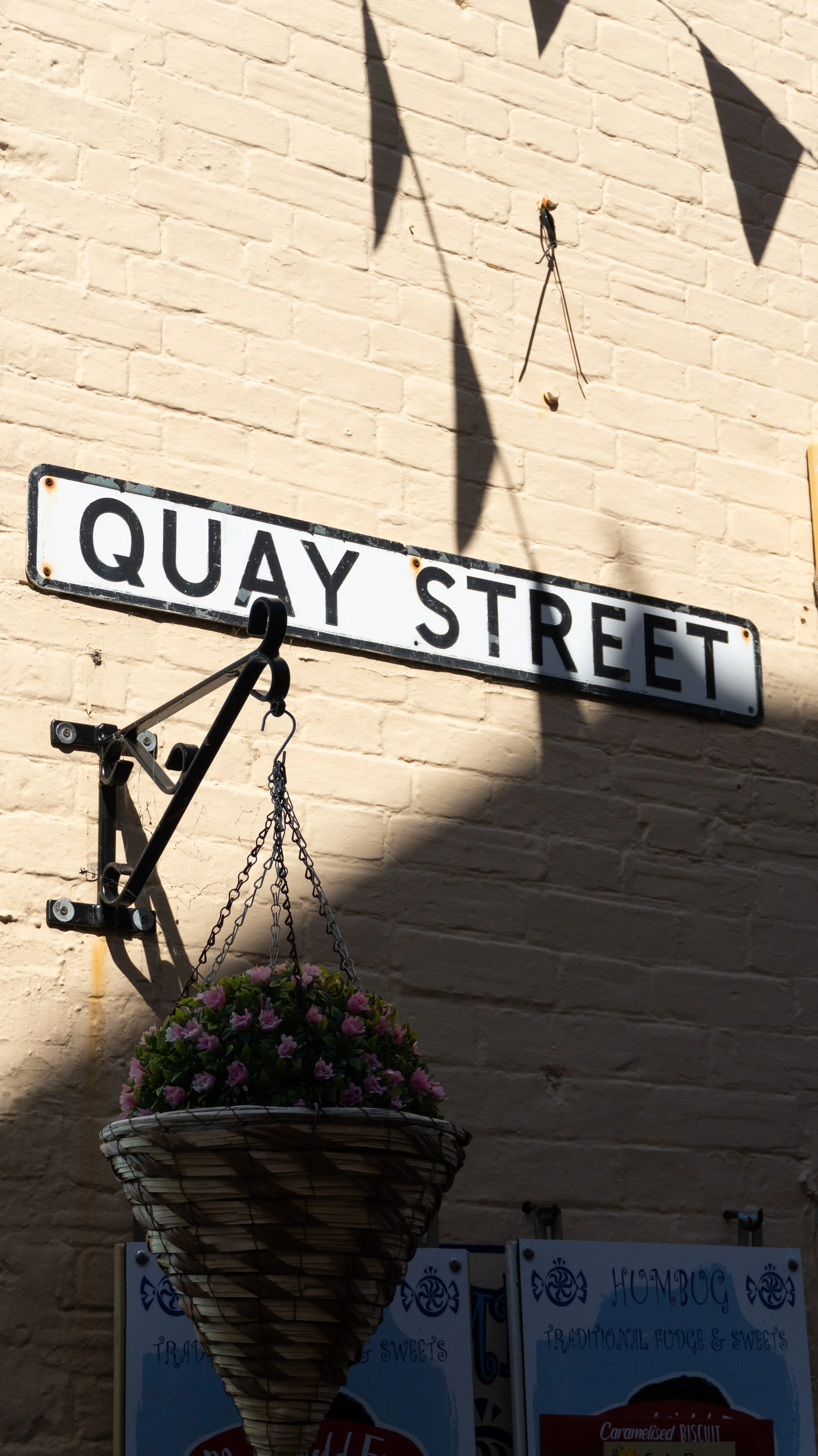 A street sign reading 'Quay Street' mounted on a beige brick wall in Lymington, with a hanging flower basket below and shadows cast on the wall.